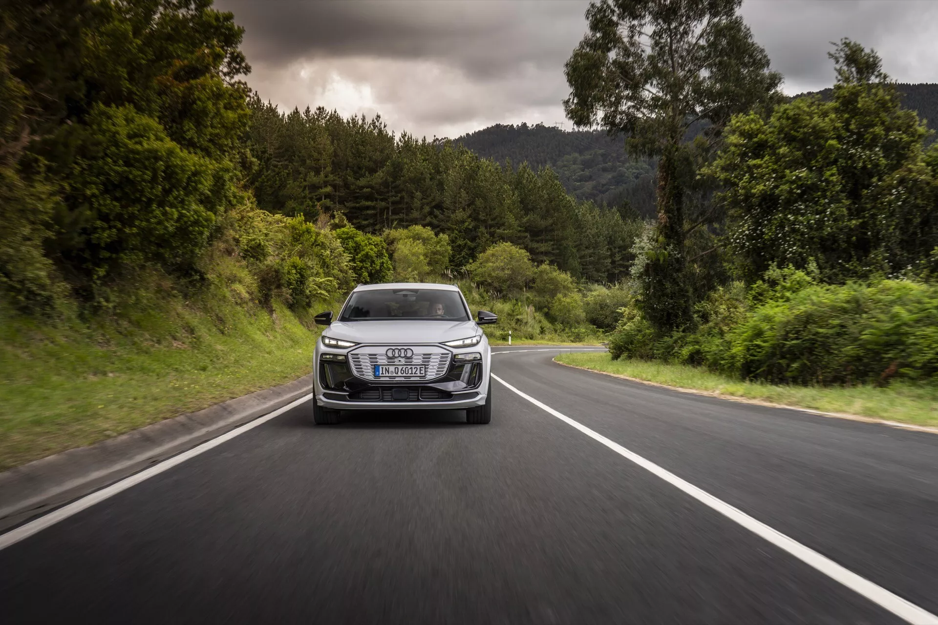 Audi Q6-e-tron 2024 ujet v gibanju iz sprednjega kota na dvopasovni cesti, obdani z zelenjem. Fotografija prikazuje rešetko, žaromete, pokrov motorja in sprednji odbijač vozila, poudarja njegovo dinamično zmogljivost in oblikovanje. Avto ima značilne LED žaromete, izrazit luksuzni emblem na rešetki in aerodinamične črte, vse v oblačnem vremenu z suhimi cestnimi pogoji.