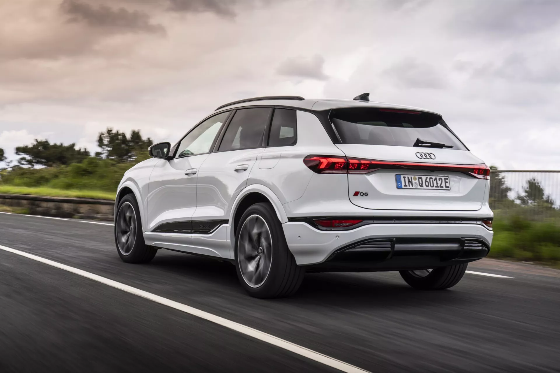 Audi Q4 e-tron 2024 v gibanju na asfaltirani cesti, prikazan iz zadnjega trikratnega pogleda, ki zajema zadnji levi del. Slika prikazuje tanke zadnje luči, strešni spojler, panoramsko streho, večkrake aluminijaste platišča, dvobarvno karoserijo z kovinskimi obrobami in ikonski znak Audi. Zamegljenost koles in zelena okolica ob cesti z oblačnim nebom nakazujeta dinamične pogoje vožnje v vseh vremenskih pogojih.