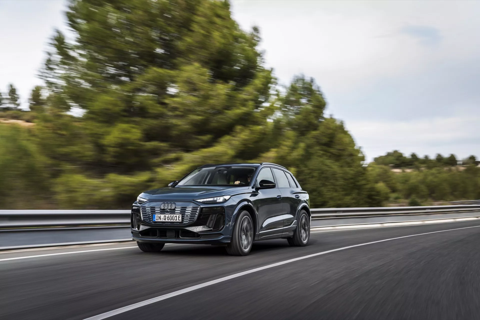 Audi Q6 e-tron 2024 SUV v gibanju na avtocesti, zajet iz trikratrinskega sprednjega pogleda. Slika prikazuje sprednjo rešetko, LED žaromete, pokrov motorja, vetrobransko steklo in del bočnega profila, vključno z vrati in stranskimi ogledali. Vozilo vozi po desni strani pasu, pomika se z leve proti desni z zamegljenim ozadjem, kar nakazuje hitrost. Drevesa in varnostna ograja so rahlo vidni. Jasna zasnova sprednjih žarometov nakazuje napredne sisteme razsvetljave, velika rešetka pa nakazuje tehnologijo hlajenja ali senzorje za ADAS. Stranska ogledala lahko vsebujejo smerokaze ali sisteme za spremljanje mrtvih kotov. Kolesa dajejo vpogled v zmogljivostne ali luksuzne kategorije.