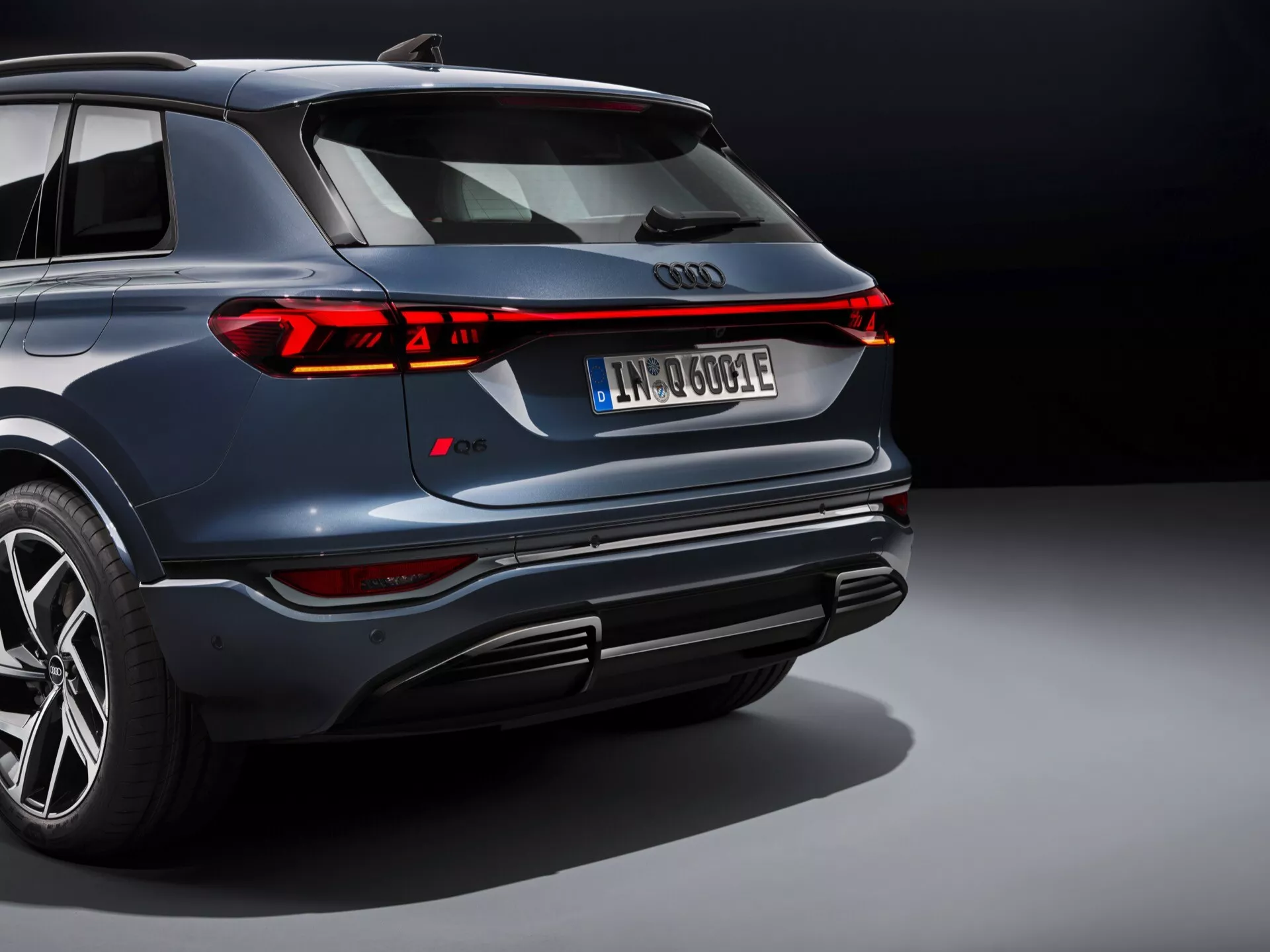 Ta slika prikazuje sodoben SUV, verjetno Audi Q6 e-tron 2024, gledan iz dvignjenega zadnjega kota. Vidimo ikoničen štirirep Audi emblem na hrbtni strani, obdan z elegantnim neprekinjenim LED trakom zadnjih luči, ki se razteza čez celotno širino vozila, s segmentiranimi zavorami in smerniki. Temno mornarsko modro vozilo ima svetleče kovinsko obdelavo, drzno oblikovanje odbijača z integriranim izpušnim sistemom in difuzorjem, večkrakimi litoželeznimi kolesi z nizkoprofilnimi pnevmatikami ter registrsko tablico, ki nakazuje električni ali hibridni pogonski sklop. Profesionalna osvetlitev in nevtralno ozadje poudarjata luksuzen in napreden dizajn vozila, pri čemer izpostavljata njegove športne in visoko zmogljive lastnosti.