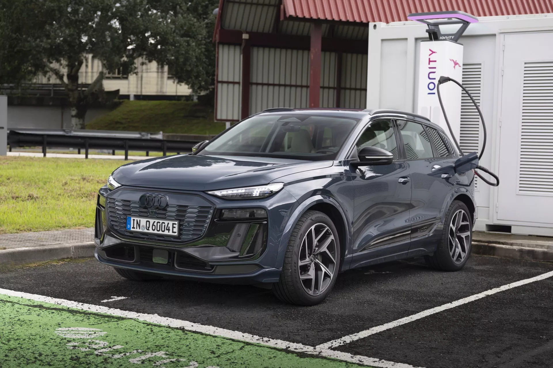 Slika prikazuje sodoben SUV Audi Q6 e-Tron 2024 parkiran pri polnilni postaji za električna vozila. Avto je prikazan iz rahlo kotnega sprednjega pogleda, poudarjajoč njegovo masko, žaromete in del pokrova motorja ter vetrobranskega stekla. Položen je diagonalno, s sprednjim delom rahlo obrnjenim na levo. Vidna je leva stran vozila, vključno s sprednjim kolesom na voznikovi strani, vrati in stranskim ogledalom. SUV je priključen na polnilno postajo na levi sprednji strani nad kolesom. Avto ima elegantne linije, zaprto masko, značilno za električna vozila, in napredne LED žaromete. Podrobnosti nakazujejo možne napredne sisteme za pomoč vozniku, kot so prilagodljivi tempomat, pomoč za ohranjanje voznega pasu in sistem za preprečevanje trkov. Slika je svetla in jasna, posneta podnevi brez zameglitve zaradi gibanja, tako da so vsi oblikovni in tehnološki elementi lahko vidni.