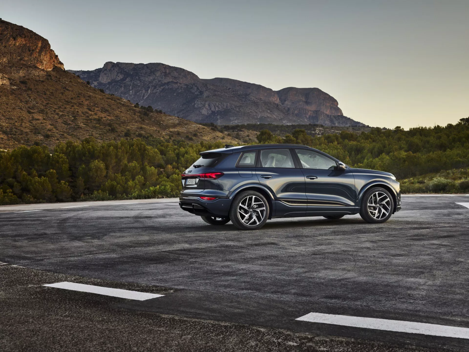 Slika SUV vozila Audi Q6-e-tron 2024, parkiranega na zunanjem parkirišču s naravno, morda gorato pokrajino v ozadju. Fotografija zajema pogled s treh četrtin od zadaj, poudarja oblikovne elemente bočne in zadnje stranice. Vidna je voznikova stran, ki prikazuje oblikovne linije, zadnja okna in oblikovanje koles. Nagnjena strešna linija nakazuje na športen, kupejevski dizajn SUV-ja. Moderna LED zadnja luč, vidni senzorji ali odsevniki na zadnjem odbijaču ter dvobarvna barvna shema poudarjajo višino in držo. Velika aluminijasta platišča in obsežne pnevmatike kažejo na višjo opremo s športno estetiko. Vozilo je videti blizu kamere, kar ponuja podroben pogled. Pomembne značilnosti vključujejo LED osvetlitev, parkirne senzorje, visokozmogljiva kolesa in pnevmatike, aerodinamičen dizajn ter verjetno napredne sisteme za pomoč pri vožnji in štirikolesni pogon.