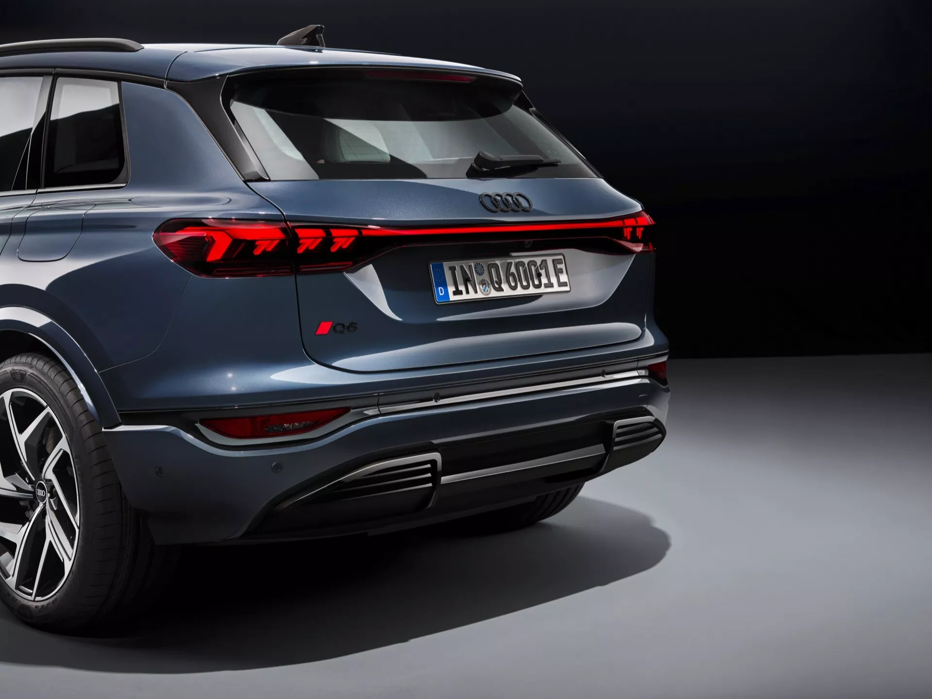 Zadnji kotni pogled na Audi Q6 e-tron 2024, z elegantnimi vodoravnimi LED zadnjimi lučmi, osrednjim logotipom Audi, značko 'e-tron', ki označuje električni model, evropsko registrsko tablico 'IN 600 E', športnim zadnjim odbijačem s ponarejenimi izpušnimi cevmi, simetričnimi odsevnimi elementi in lučmi na skrajnih delih odbijača, temno modrim kovinskim lakom, ki odseva studijsko osvetlitev, delno vidnim športnim desnim zadnjim kolesom in enotnim temnim studijskim ozadjem.