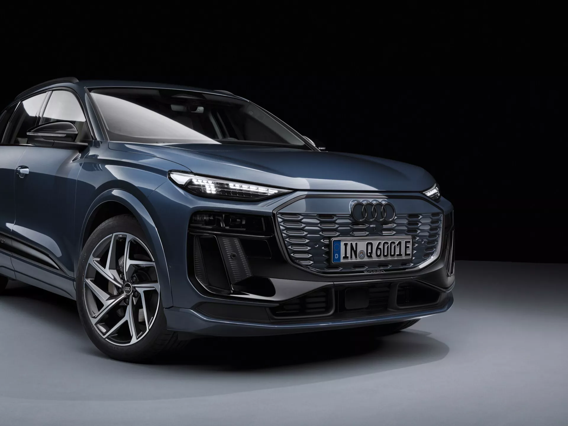 Slika prikazuje sodobni Audi Q6 e-tron 2024, posnet iz nizkega prednjega trikotnega kota. Vidna je ikonična sprednja maska z vodoravnimi letvicami, zapleteni LED žarometi, agresivni sprednji odbijač z velikimi dovodi zraka, večkraki aluminijasti platišči in značka elektrificiranega modela. Elegantni in izpopolnjeni SUV prikazuje aerodinamične linije in napredno zasnovo na profesionalni studijski fotografiji proti črnemu ozadju.