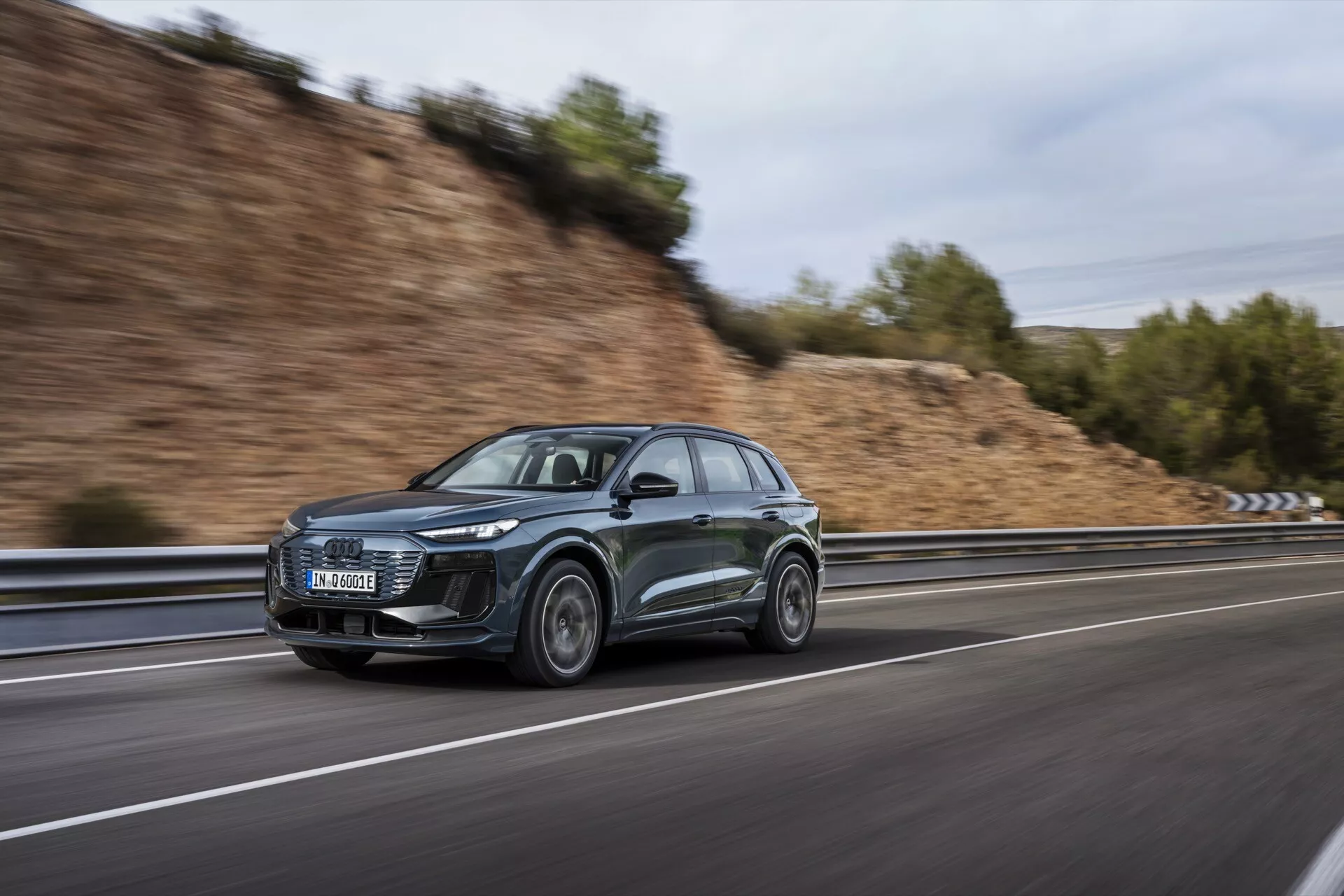 Ta slika prikazuje eleganten in prefinjen SUV Audi Q6 e-tron 2024, posnet iz stranskega kota v gibanju. Sodobno zasnovo vozila poudarjajo izraziti LED žarometi in aerodinamične lastnosti, vključno z nagnjeno strešno linijo in aerodinamičnimi stranskimi ogledali. Avto je končan v temni barvi, verjetno sivi ali črni, in se vozi po hitri cesti, kar dokazuje zamegljeno ozadje, ustvarjeno s tehniko panoramske fotografije. Prednje kolo je rahlo zasukano proti kameri, kar prikazuje zasnovo zlitinskega kolesa. Dodatni vidni elementi vključujejo možno vgrajeno tehnologijo v žarometih in stranskih ogledalih ter potencialne strešne nosilce, osredotočene na uporabnost.
