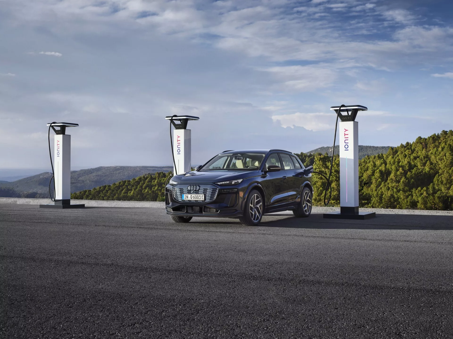 SUV Audi Q6 e-tron 2024 v globoko kovinsko modri barvi na polnilni postaji za električna vozila s tremi polnilniki. Slika zajame vozilo pod tremi četrtinskimi kotom, prikazuje njegovo prednjo levo stran, poudarja rešetko, žaromete, kolesa in pokrov motorja. Eleganten in sodoben električni avtomobil ima športno in vsestransko zasnovo z elegantnimi, agresivnimi žarometi in platišči iz lahke litine, poudarja svojo napredno, trajnostno avtomobilsko tehnologijo.