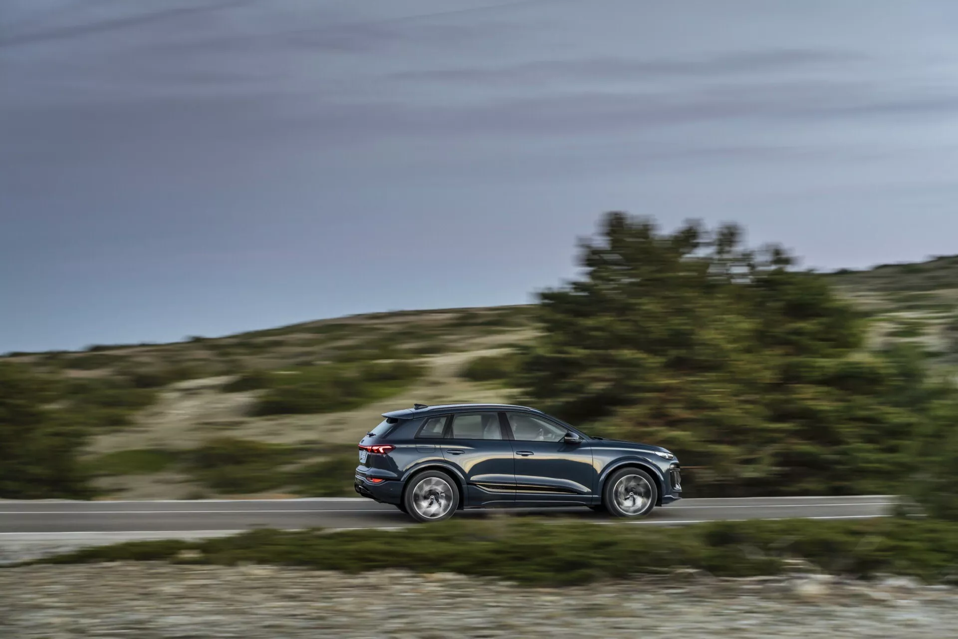 Dinamičen stranski pogled na Audi Q6 e-tron 2024 v gibanju z zamegljenim ozadjem, srednji paralelni kot prikazuje profil, oblikovalske linije, okna, pet vrat, stranska ogledala, platišča iz zlitine, dnevna nastavitev, podeželsko ozadje.