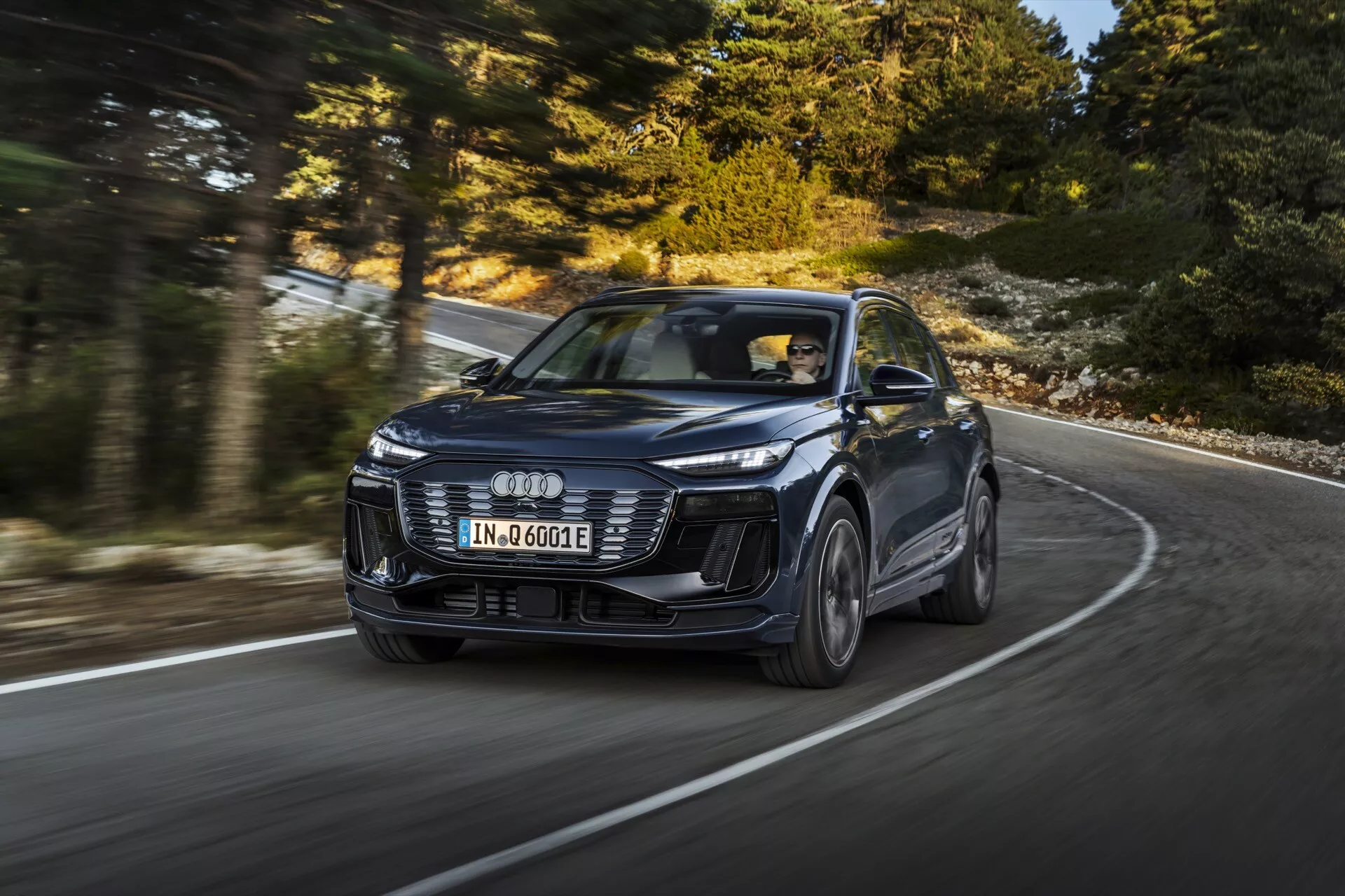 Slika moderne SUV Audi Q6 e-tron 2024 v gibanju, posneta iz dinamičnega tričetrtinskega sprednjega kota, ki poudarja oblikovanje in zmogljivost. Vozilo, prepoznavno po značilnem logotipu s štirimi obroči, ima emblematične LED žaromete, agresiven sprednji odbijač in oblikovane črte pokrova motorja. Slika, posneta iz nižjega kota, poudarja prisotnost avtomobila na zaviti cesti, pri čemer so platišča rahlo zamegljena, kar kaže na gibanje. Novi model nakazuje napredno tehnologijo, kot je prilagodljiv tempomat in električni pogon, označen z oznako 'e-tron'. Profesionalno fotografiranje, ki prikazuje razkošje, oblikovanje in tehnologijo v konfiguraciji z vožnjo na levi strani.