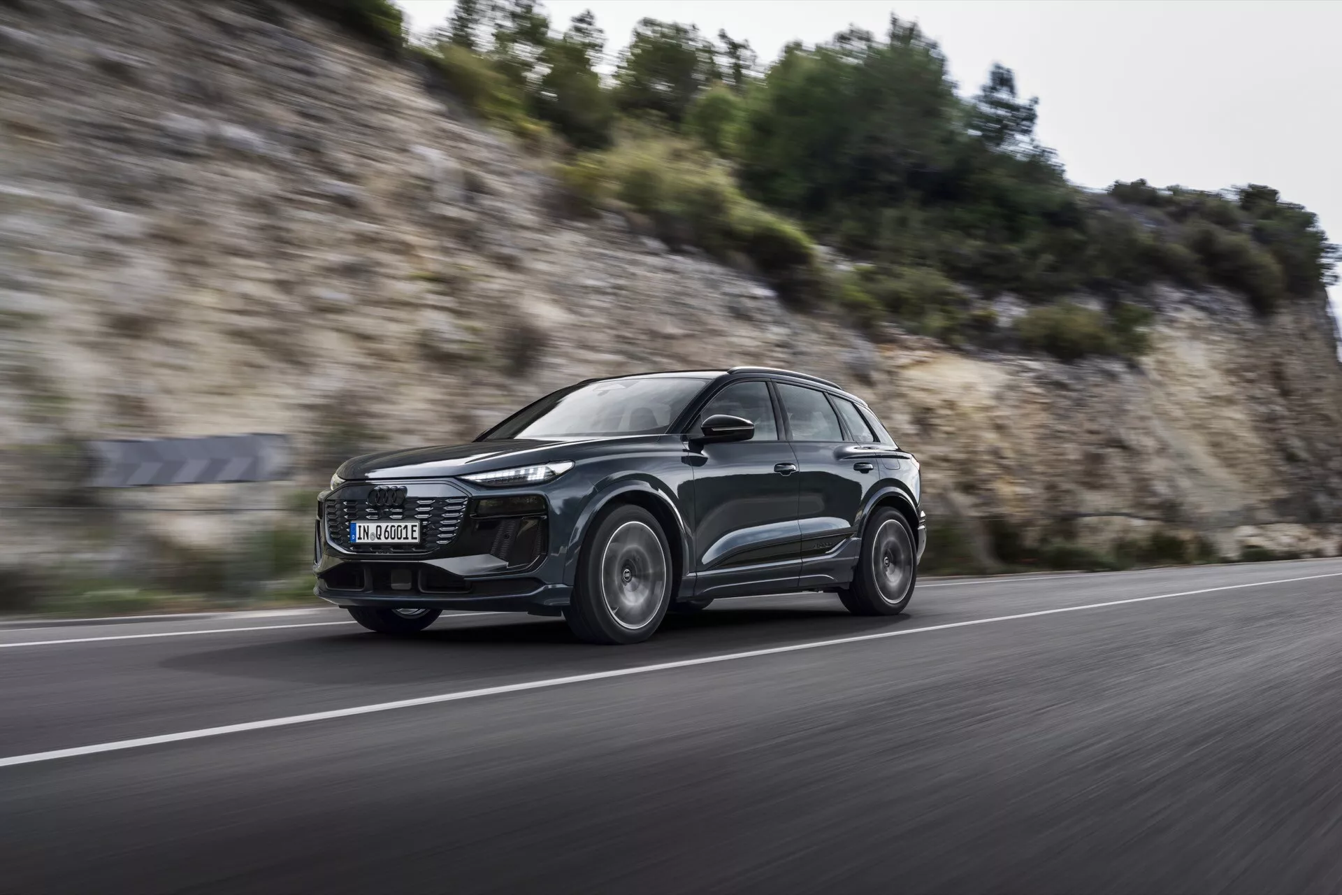 Dinamičen posnetek Audi Q6 e-tron 2024 v gibanju iz nizkega sprednjega kota, ki prikazuje njegov eleganten aerodinamičen SUV dizajn, moderne LED žaromete in zlitine koles s zameglitvijo gibanja, kar nakazuje zmogljivostne sposobnosti na asfaltni cesti.