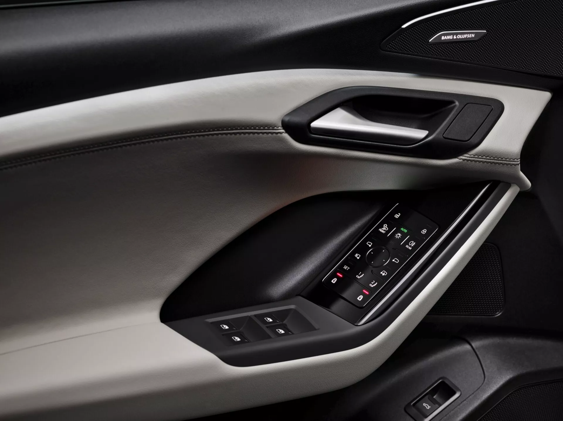 Pogled od blizu na notranjo ploščo vrat Audi Q6 e-tron 2024, ki poudarja oblikovne elemente in funkcije. Plošča vrat, posneta iz diagonalnega kota, razkriva različne materiale in teksture, vključno z zgornjim delom, mehkim na dotik, z vidnimi šivi, trdim oblogom, ki vsebuje ergonomsko ročico, in oblazinjenim naslonom za roke s svetlečo upravljalno ploščo za okna in nastavitev stranskih ogledal. Pod naslonom za roke je zvočni sistem z znamko, kar kaže na vrhunski avdio sistem. Barvna shema vključuje dvobarvno obliko, ki povečuje estetske in funkcionalne lastnosti.