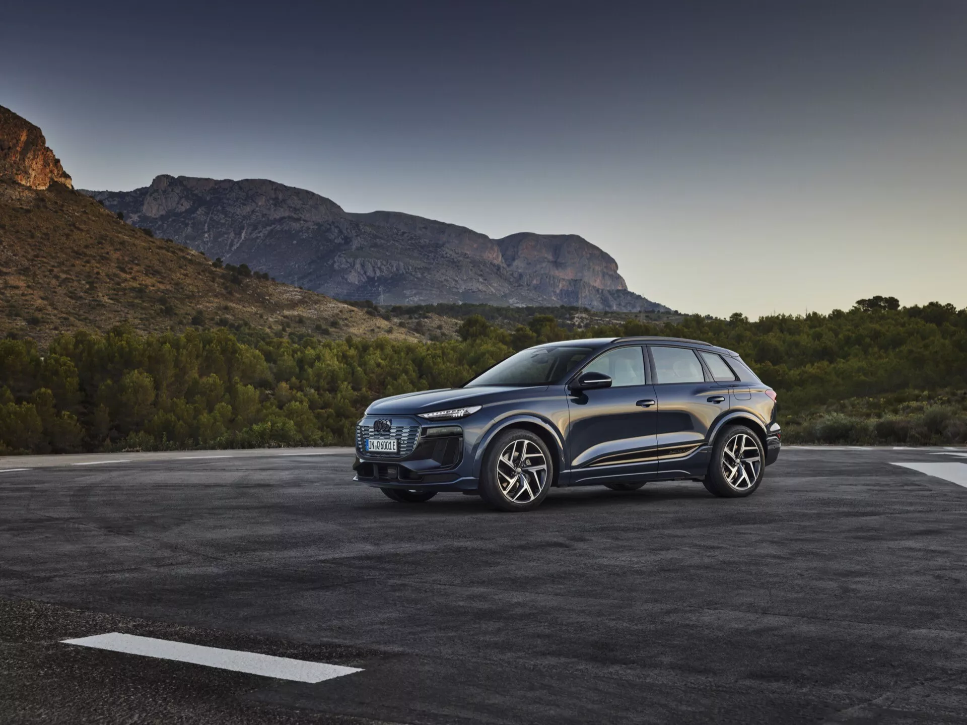 Trikratni sprednji pogled na modri SUV Audi Q6 e-tron 2024, parkiran na prostem, ki prikazuje izrazito mrežo, elegantne žaromete, oblikovano pokrov motorja, velika lita platišča, stranska ogledala in voznikova vrata ob slikovitem gorskem ozadju v zgodnjem jutranjem ali pozno popoldanskem svetlobi.