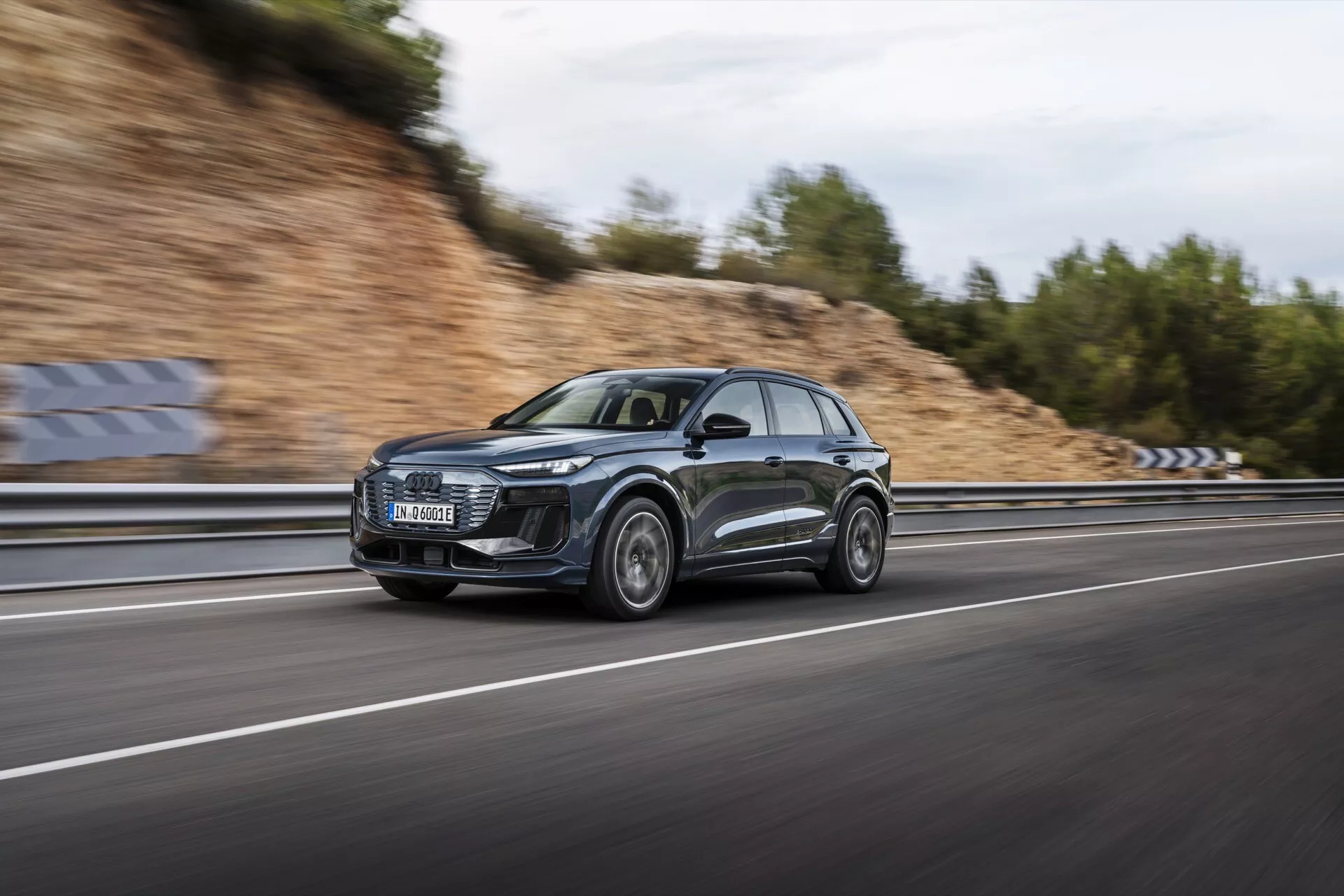 Slika Audi Q6 e-tron 2024 v gibanju, posneta s tehniko panoramskega snemanja, poudarja eleganten profil in sodoben dizajn SUV-ja. Avto, posnet s strani, se hitro vozi po večpasovni avtocesti z zamegljenimi kolesi in ozadjem, kar poudarja njegove dinamične zmožnosti. Temno kovinsko vozilo ima veliko sprednjo masko, značilne LED luči, športna aluminijasta platišča in odsevne površine. Oblačno nebo in mokra cesta ustvarjata dramatično sceno, ki poudarja luksuzne in zmogljivostne lastnosti avtomobila.