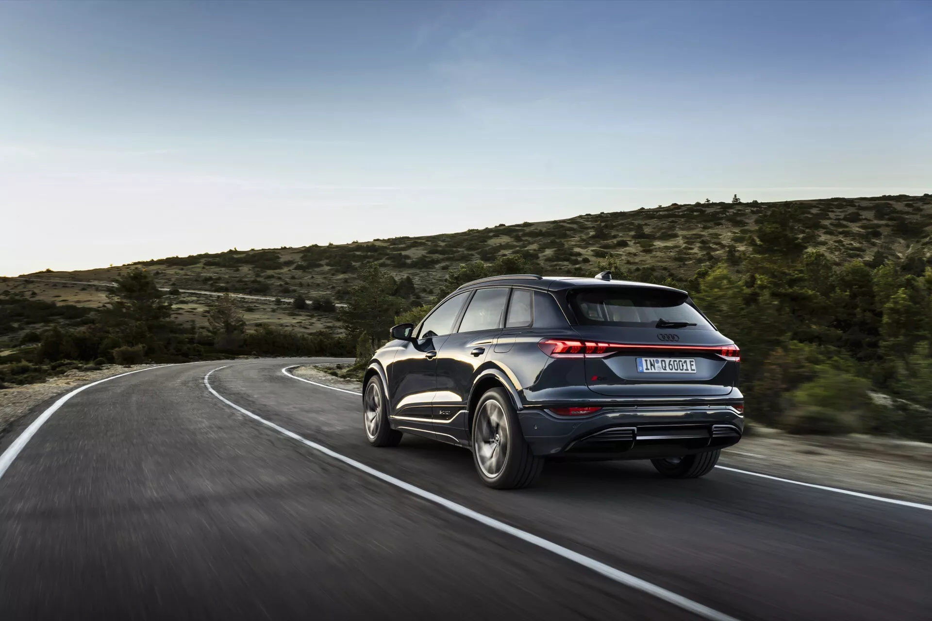 Slika prikazuje Audi Q6 e-tron 2024 iz zadnjega tristranskega kota, poudarjajoč desno stran in zadnji del vozila. Avto, ki se zdi, da je v gibanju na vijugasti cesti, prikazuje svoje oblikovalske elemente, vključno z zadnjimi lučmi, zadnjim odbijačem, izpušnimi odprtinami, aluminijastimi platišči in delom stranskega profila z zadnjimi potniškimi vrati in oknom. Zamegljeno ozadje nakazuje hitrost, naravna osvetlitev pa nakazuje, da je bila fotografija posneta podnevi v podeželskem ali polpodeželskem okolju z redko vegetacijo. Predlagani so napredni elementi, kot so LED zadnje luči, zadnji spojler ali dvojni izpuhi, čeprav so drobnejše tehnološke podrobnosti zaradi zamegljenosti gibanja neprepoznavne.