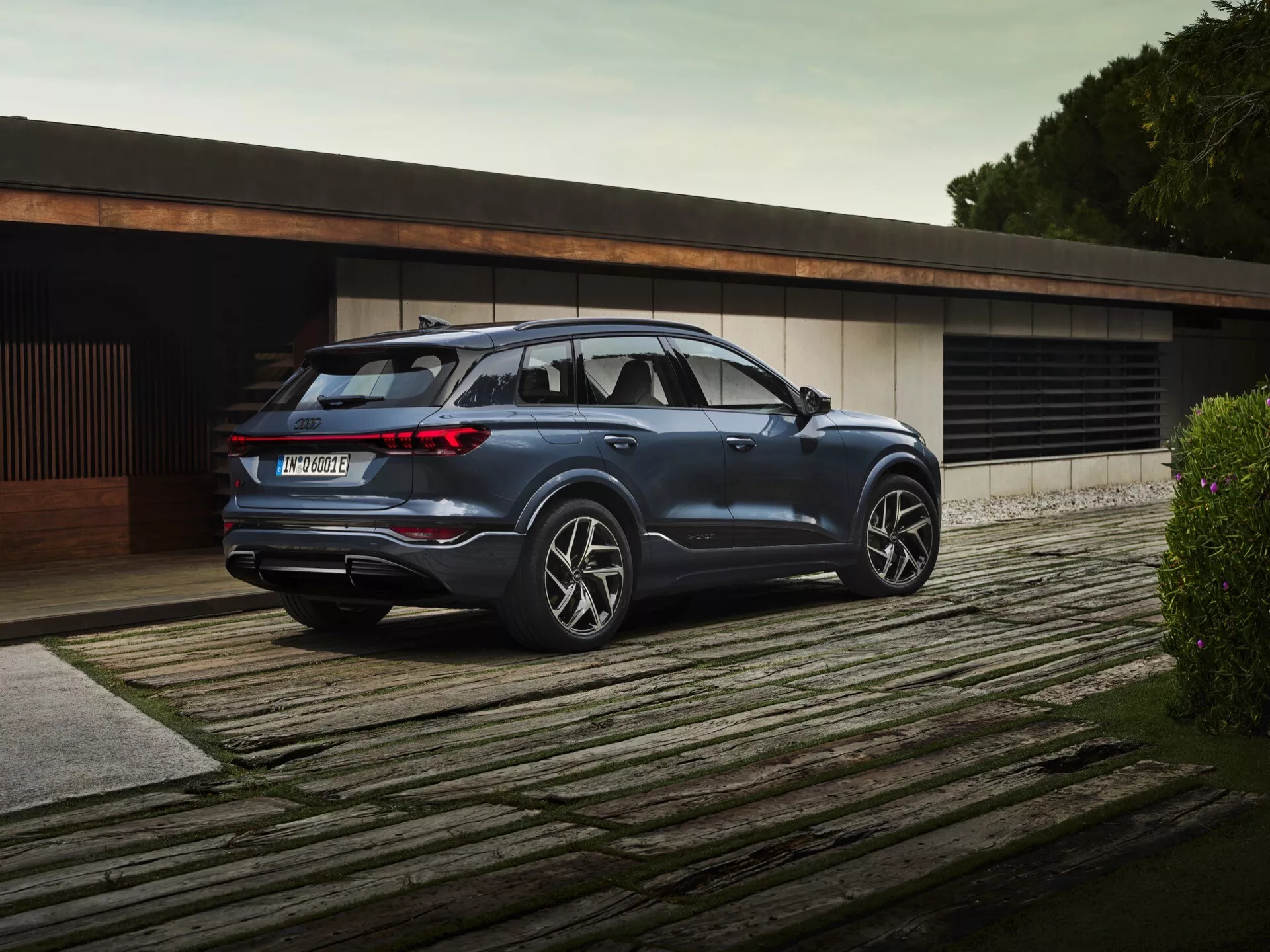 Slika modela Audi Q6-e-Tron 2024, parkiranega na leseni ploščadi, prikazuje pogled s treh četrtin zadaj. Vidni so bočni profil in zadnji del luksuznega SUV-a, z modernimi vodoravnimi LED zadnjimi lučmi, robustnimi zadnjimi vrati, izrazitim zadnjim odbijačem z možnimi parkirnimi senzorji in difuzorjem spodaj z dvojnimi izpušnimi cevmi. Bočni profil poudarja velika večkraka platišča iz lahke litine, izrazite karakterne linije, strešne nosilce in rahlo zatemnjena zadnja okna. Tehnološki dodatki, kot so stranska ogledala z vgrajenimi smerniki in vgrajeni ročaji vrat, nakazujejo napredno, vrhunsko vozilo v sofisticiranem, minimalističnem okolju.