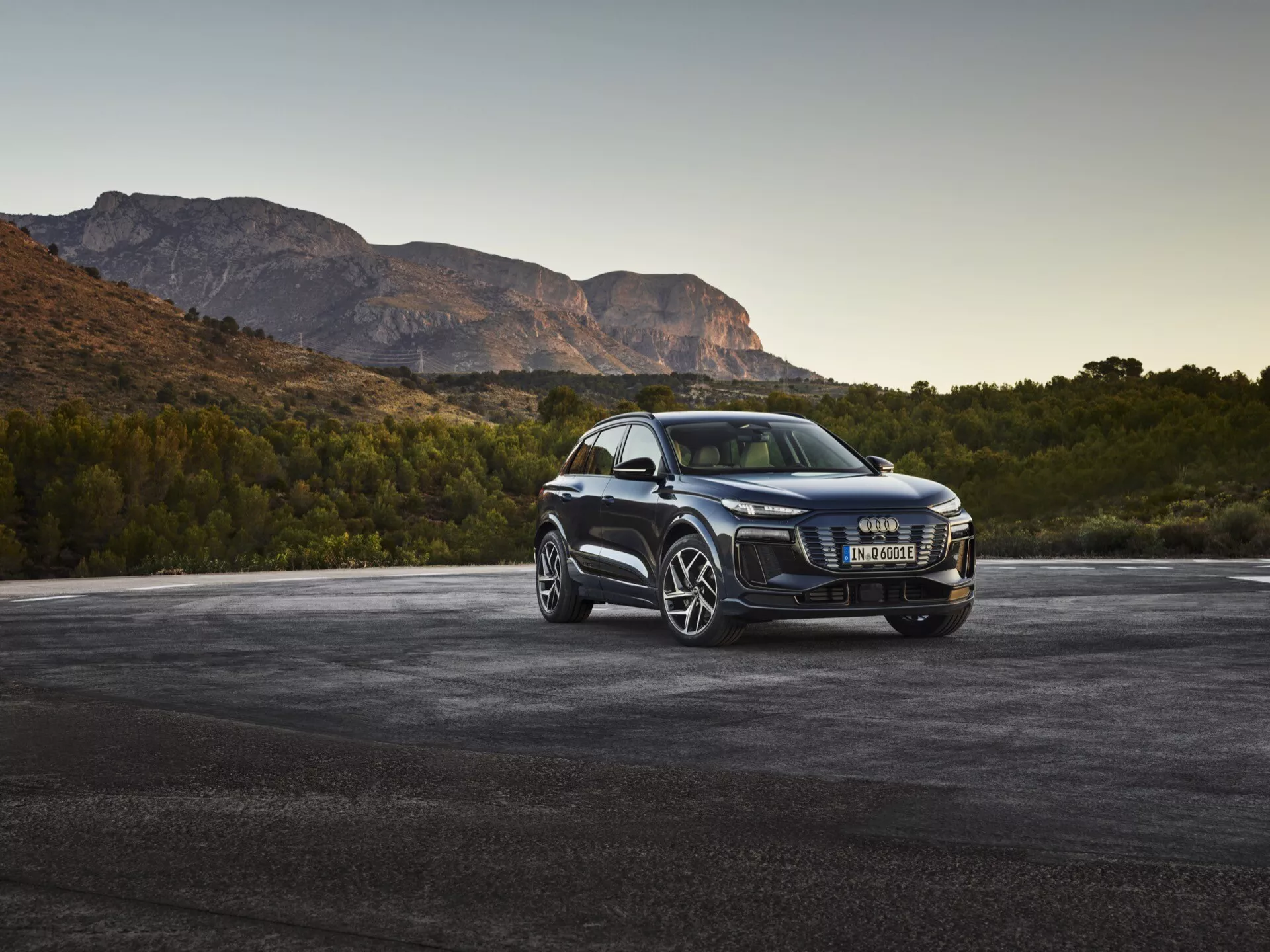 Luksuzni SUV Audi Q6 e-tron 2024 pod trikratratnim sprednjim kotom, ki prikazuje kromirano rešetko, sodobne LED žaromete, velika platišča iz več naper, stranska ogledala z vgrajenimi smerniki, zatemnjena okna in sijajno barvo, parkiran na asfaltu z gorami v ozadju ob zori ali mraku.