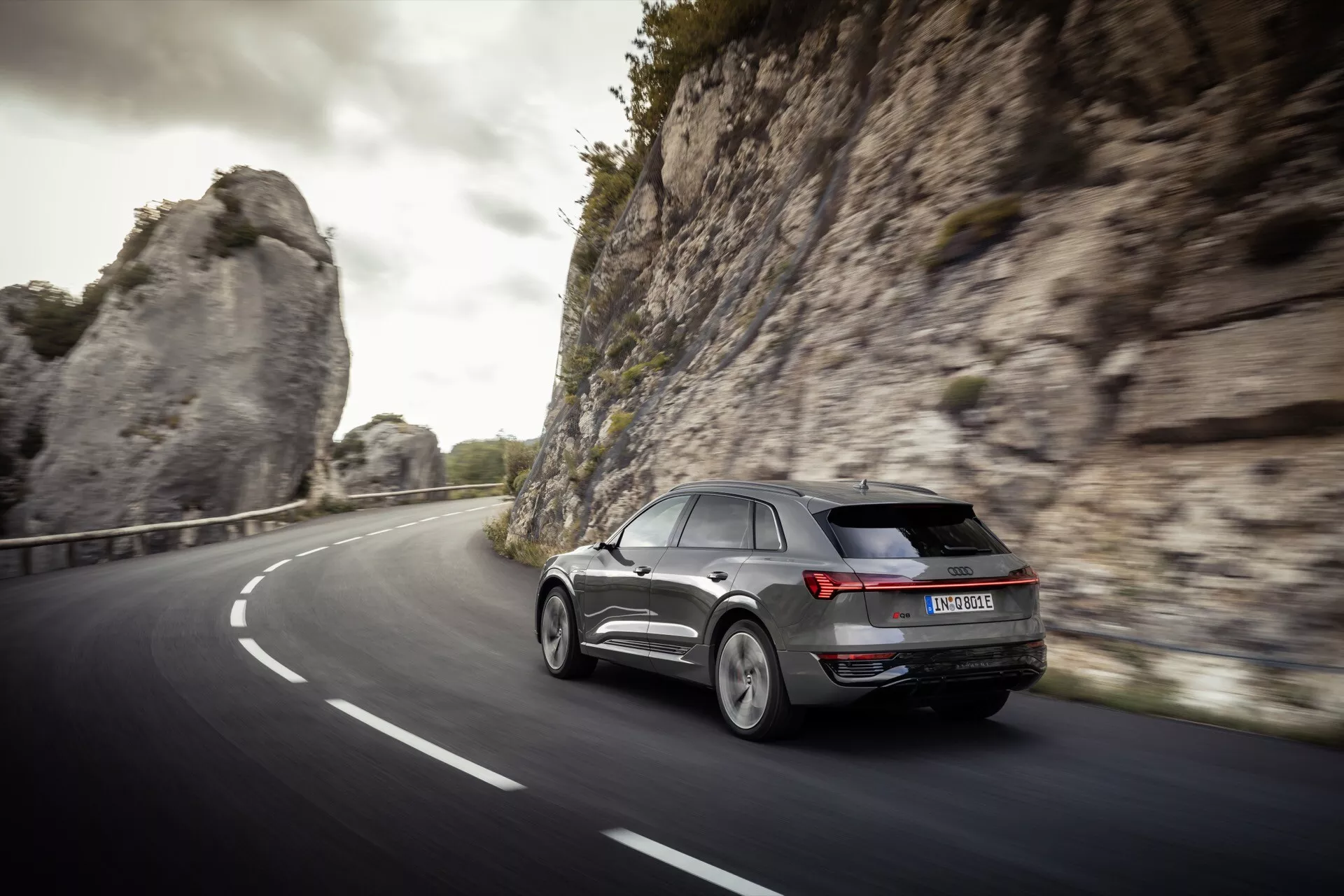 Moderen SUV Audi Q8 e-tron 2023 v dinamičnem zadnjem tričetrtinskem pogledu, vozeč po ukrivljeni dvopasovnici v naravnem, verjetno goratem okolju, ki prikazuje LED zadnje luči, lita platišča in aerodinamično obliko pod mehko svetlobo