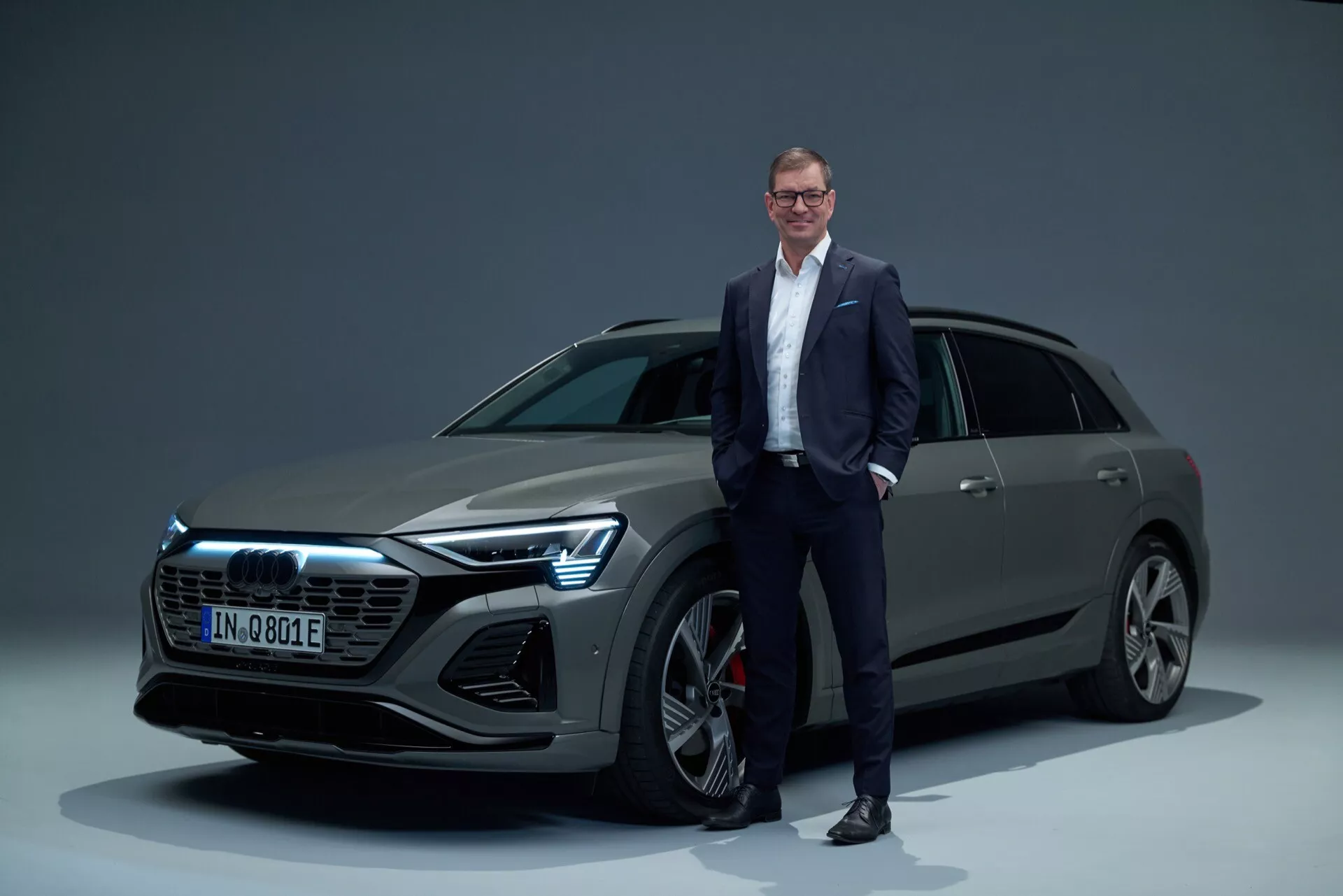 Audi Q8 e-tron 2023 v studijskem okolju, prikazuje sprednjo rešetko, LED žaromete, večkraka lita kolesa in elegantno obliko telesa SUV z moškim v poslovnih oblačilih za lestvico