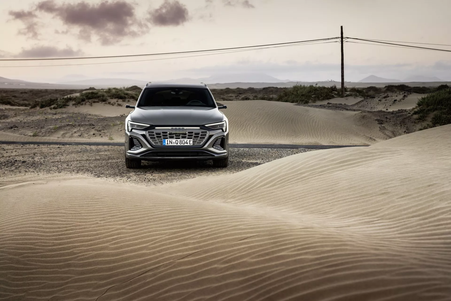 Sprednji pogled na Audi Q8 e-tron 2023 v puščavski pokrajini ob zori ali mraku, poudarjajoč njegov žar, pokrov motorja in napredno zasnovo žarometov