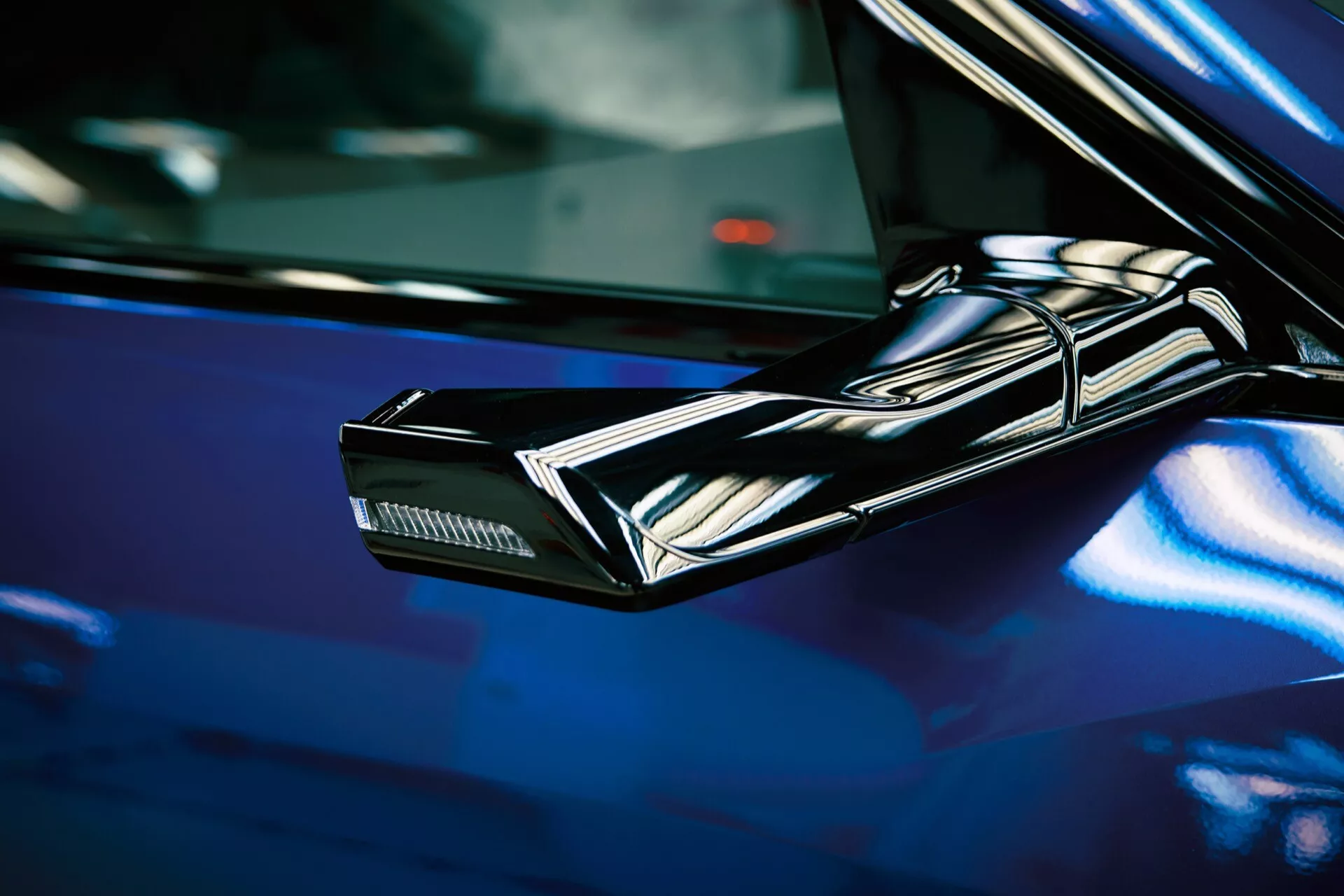 Od blizu posnet črn, eleganten, kotni stranski ogledalo Audija Q8 e-tron 2023, ki poudarja futuristične oblikovne elemente in integrirane smerne luči na ozadju globoko modre, sijoče površine avtomobila
