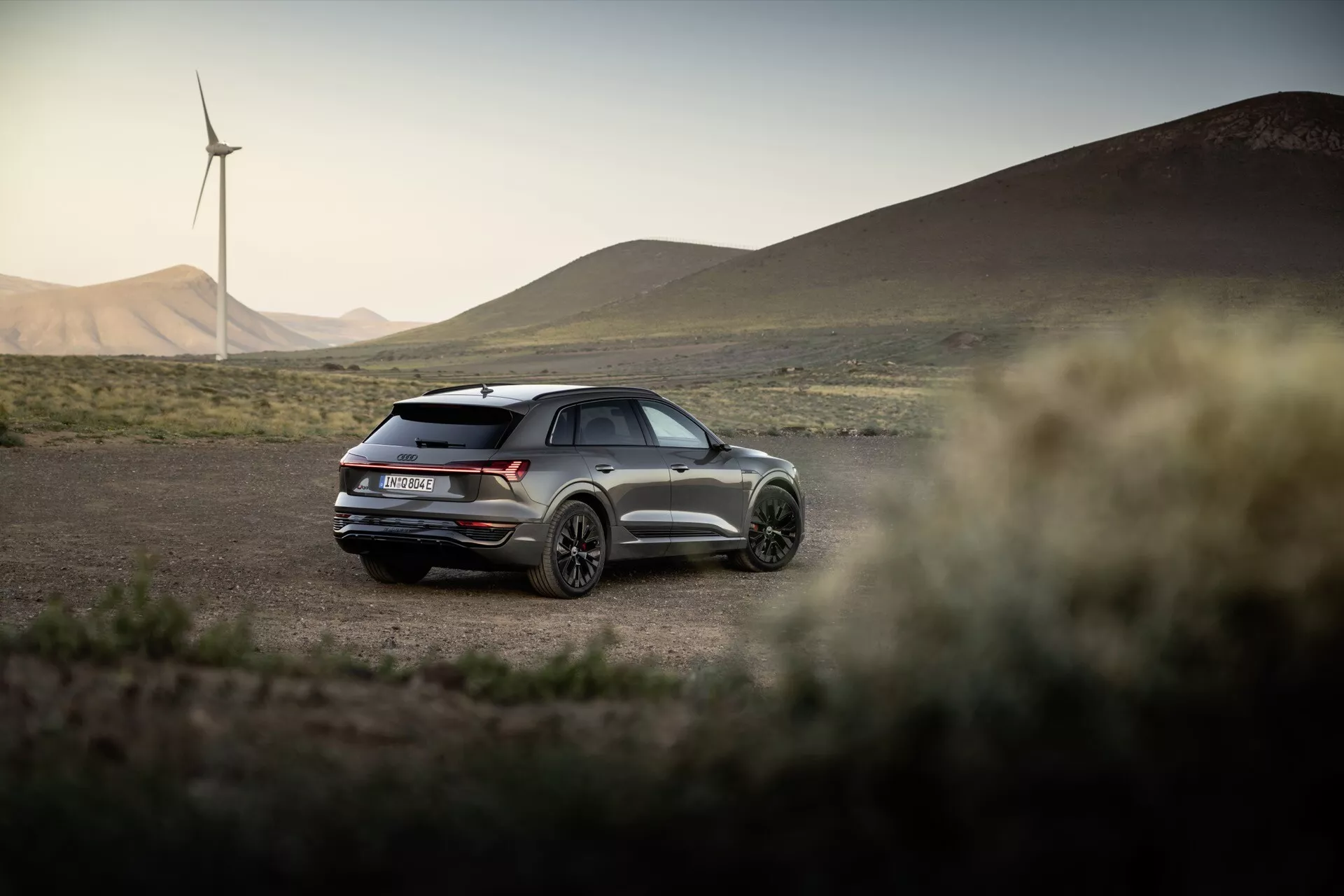 Audi Q8 e-tron 2023 v naravnem zunanjem okolju z vetrnicami v ozadju, ki prikazuje njegov športni dizajn s panoramsko streho, dvojnimi izpuhi in s tričetrtinskim pogledom od zadaj v poznem popoldnevu