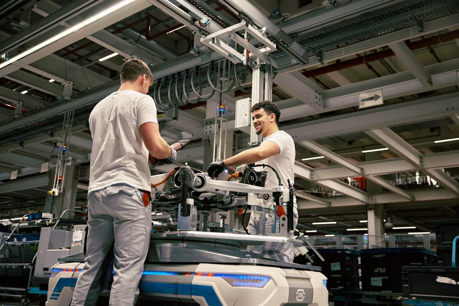 Industrijsko okolje z dvema delavcema, ki sestavljata ali vzdržujeta mehansko opremo na tleh tovarne, pod umetno razsvetljavo, z višjega vidika, ki poudarja proizvodno okolje