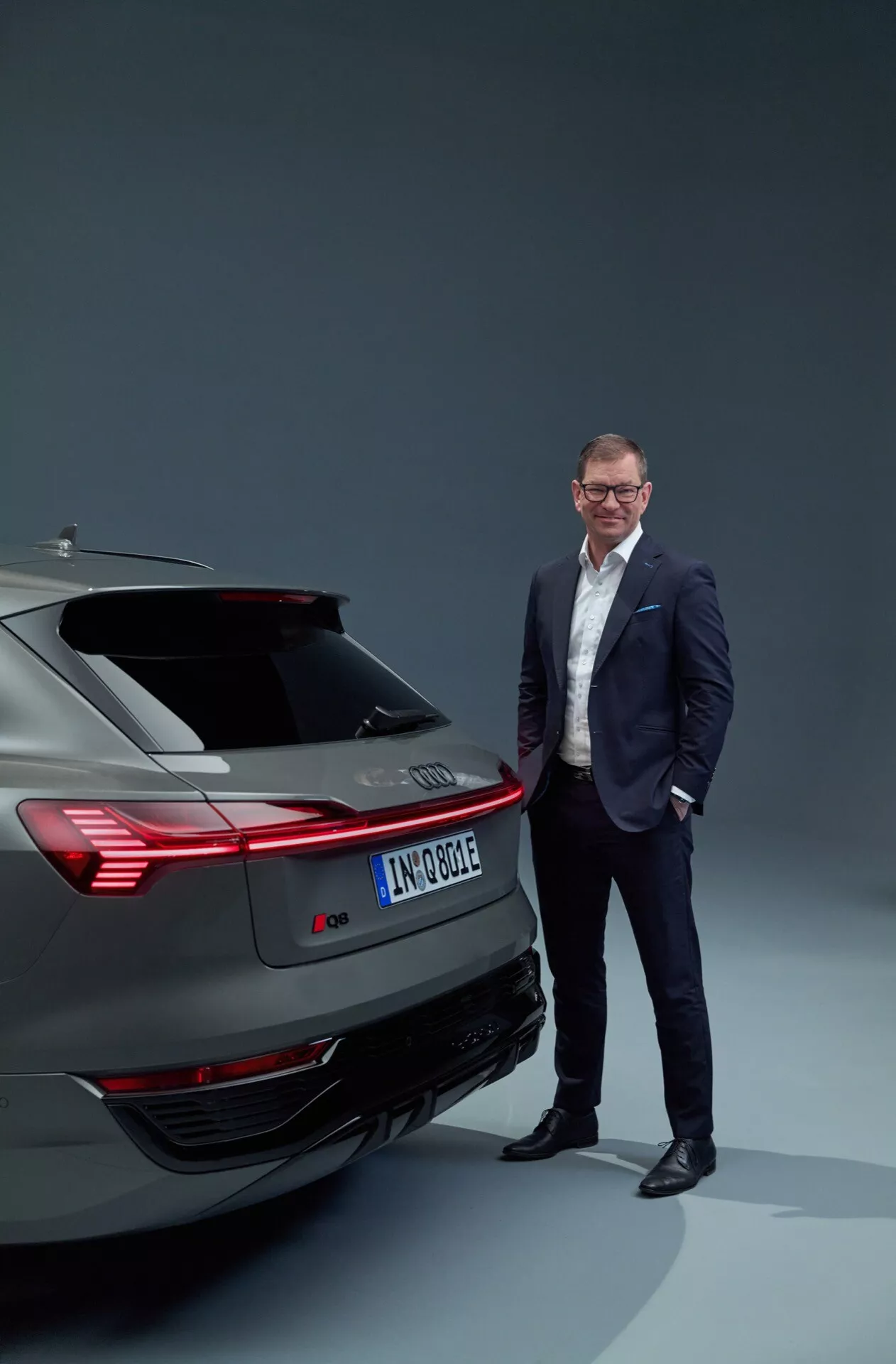 Električno vozilo Audi Q8 e-tron 2023, ki prikazuje zadnji dizajn, vključno z zadnjimi lučmi, odbijačem in izpušnim sistemom v studijskem okolju, s profesionalno oblečenim moškim, ki stoji ob strani, poudarjajoče sodobno avtomobilsko tehnologijo in luksuz.