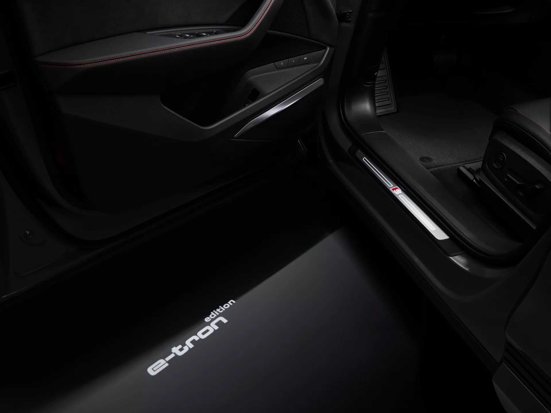 Pogled iz nizkega kota na prag vrat na strani sovoznika notranjosti Audi Q8 e-tron 2023, ki prikazuje osvetljeno napis 'e-tron', izboljšano notranjo razsvetljavo in podrobnosti premium obloge