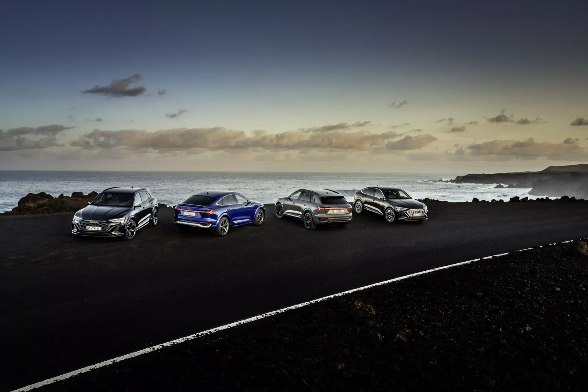 Audi Q8 e-tron 2023 parkiran na asfaltni cesti v obalnem okolju s skalnatim terenom in ozadjem oceana ob mraku, ki prikazuje aerodinamično zasnovo, zlitinska kolesa in sodobno razsvetljavo