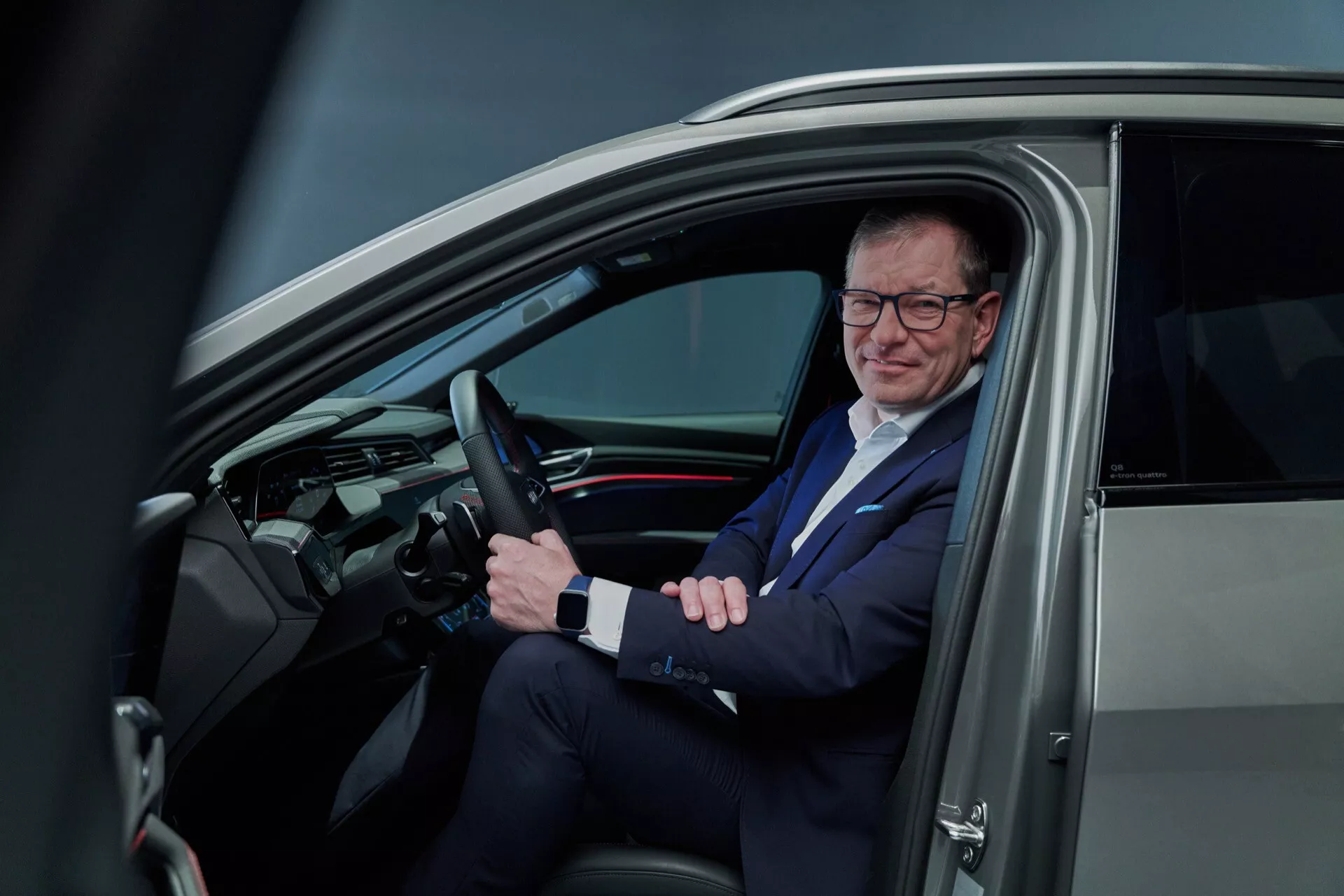 Pogled v notranjost Audija Q8 e-tron 2023 skozi odprta voznikova vrata, ki prikazuje volan z nameščenimi kontrolami, uglajeno armaturno ploščo z digitalnim zaslonom in moškega v obleki na voznikovem sedežu, kar odraža sodobno tehnologijo vozil in poslovni življenjski slog