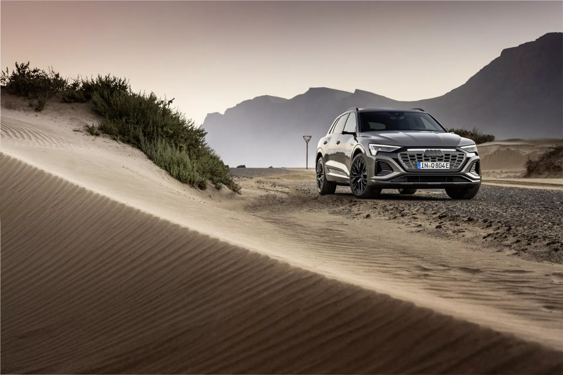 Audi Q8 e-tron 2023 v terenski nastavitvi, ki prikazuje svojo luksuzno obliko SUV, LED žaromete, zlitine kolesa in zmogljivosti delovanja proti ozadju peščenih sipin ob zlati uri