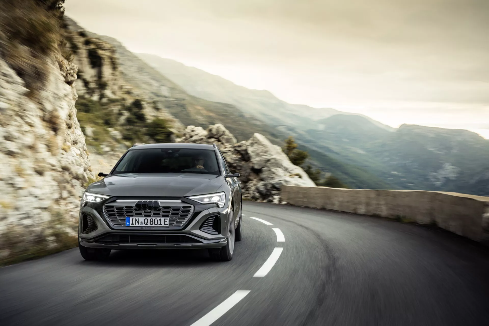Audi Q8 e-tron 2023 na vijugasti cesti z skalnatimi gorami v ozadju, ki prikazuje aerodinamično obliko in napredno tehnologijo žarometov, izraža luksuz in zmogljivost.