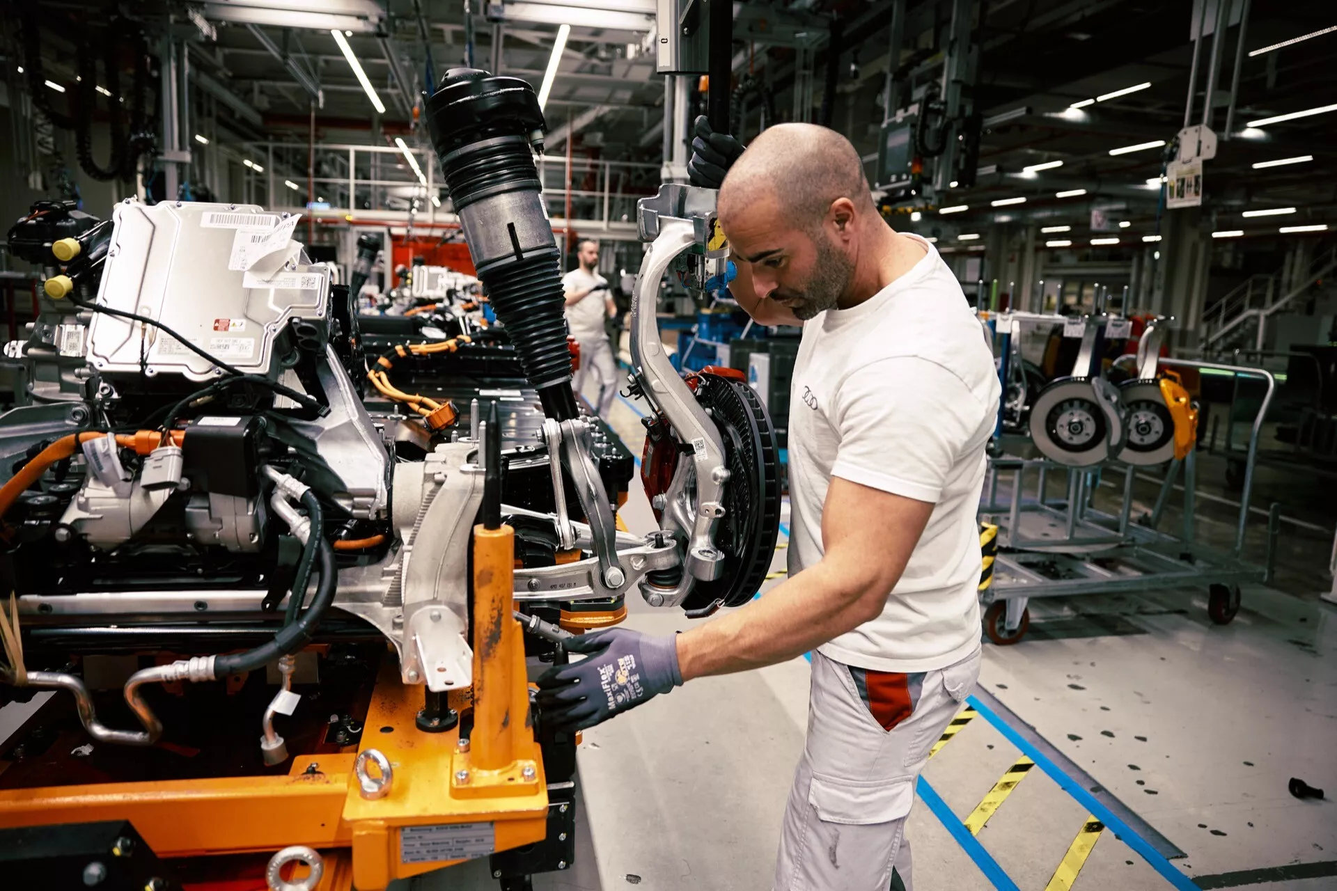 Zadnji šasija Audi Q8 e-tron 2023 na montažni stopnji z vidnimi vzmetenjem in zavornim sklopom ter delavec, ki uporablja električno orodje v obratu za proizvodnjo vozil