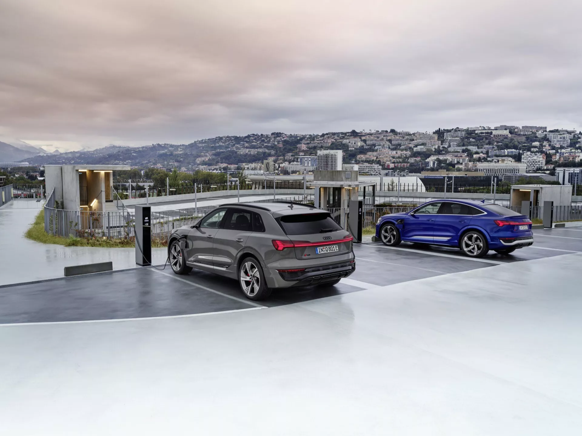 Audi Q8 e-tron 2023 v sivi in športni kupe v modri barvi parkirana na razkošnem parkirišču na strehi ob zori ali mraku z ozadjem mestne krajine, ki prikazuje napredno aerodinamiko, športno zasnovo, LED zadnje luči, dvojni izpuh in stekla za zasebnost