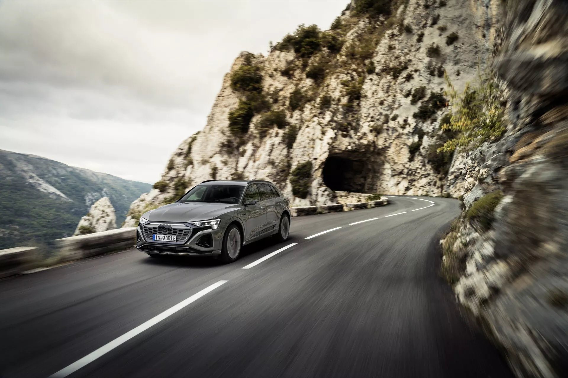 Audi Q8 e-tron 2023 dinamično se vozi po ovinkasti gorski cesti, s svojim modernim dizajnom hatchbacka, LED žarometi in veliko rešetko iz prikaza sprednjega dela na slikovitem gorskem ozadju