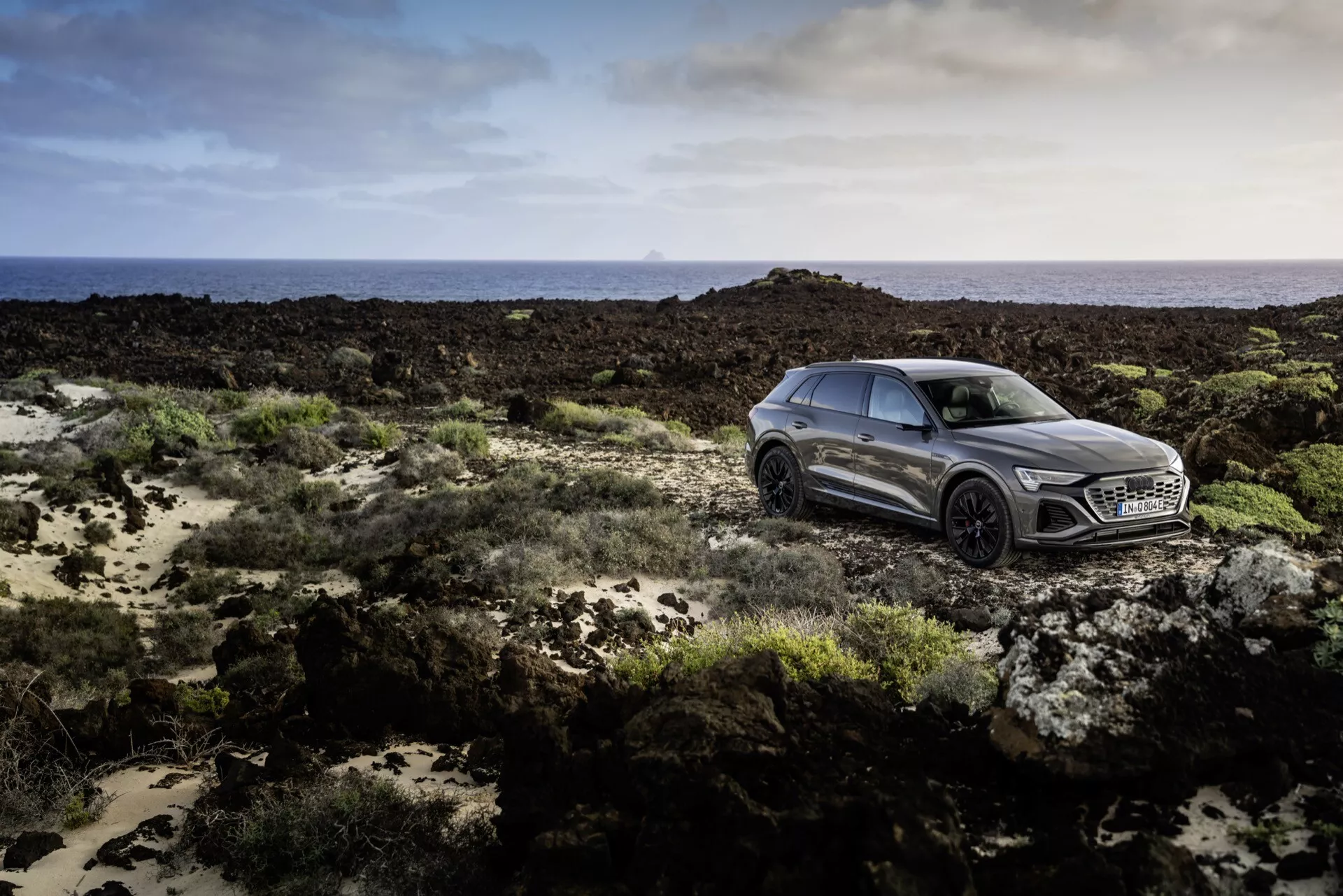 Audi Q8 e-tron 2023 v kovinski sivi barvi, ki je primer luksuza s prefinjenim, športnim dizajnom, posnet iz stranskega sprednjega kota na skalnatem terenu pod mehko osvetlitvijo, prikazuje napredno tehnologijo LED žarometov, izstopajočo masko z velikim emblemom, dinamične bočne konture in večkraka lita kolesa.