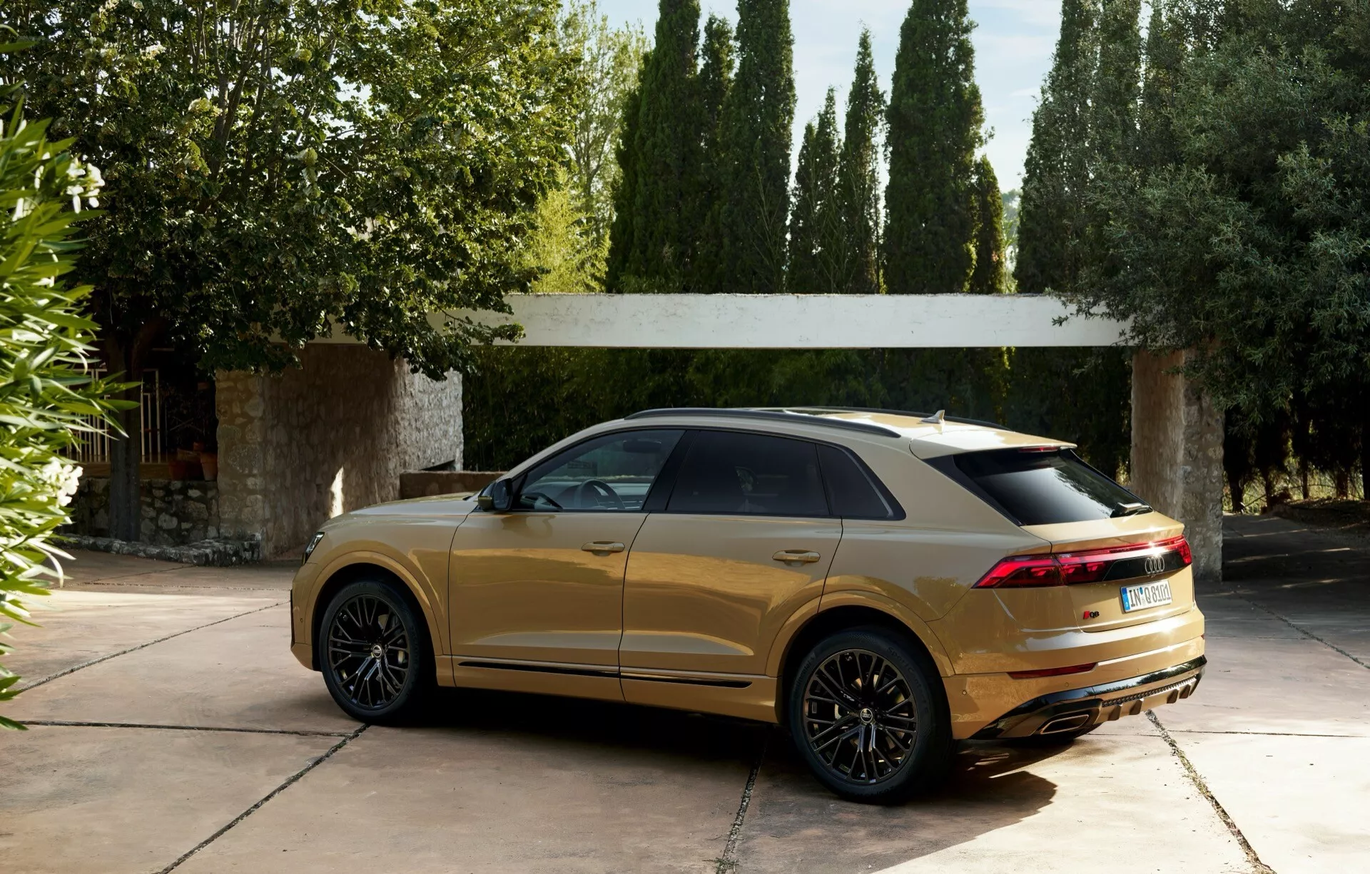 Luksuzni SUV Audi Q8 (4M-Facelift) iz leta 2023 parkiran na prestižnem dovozu, prikazuje športni zadnji spojler, elegantno padajočo streho in sodobne LED zadnje luči, v metalik zlati barvi z odsevi na sijoči karoseriji, obdan z drevesi in razkošno strukturo v ozadju
