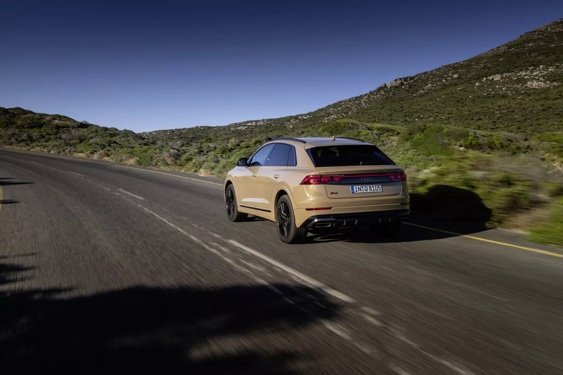 Audi Q8 2023 (4M-Facelift) v gibanju, posnet iz zadnjega tristranskega zornega kota, ki prikazuje desno stran in zadnji del na deljeni cesti z naravnim ozadjem hribov pod modrim nebom, predstavljajoč kupejevsko linijo strehe, elegantno zasnovo, podaljšane zadnje luči, svetlo bež barvo in dvojni izpušni sistem.
