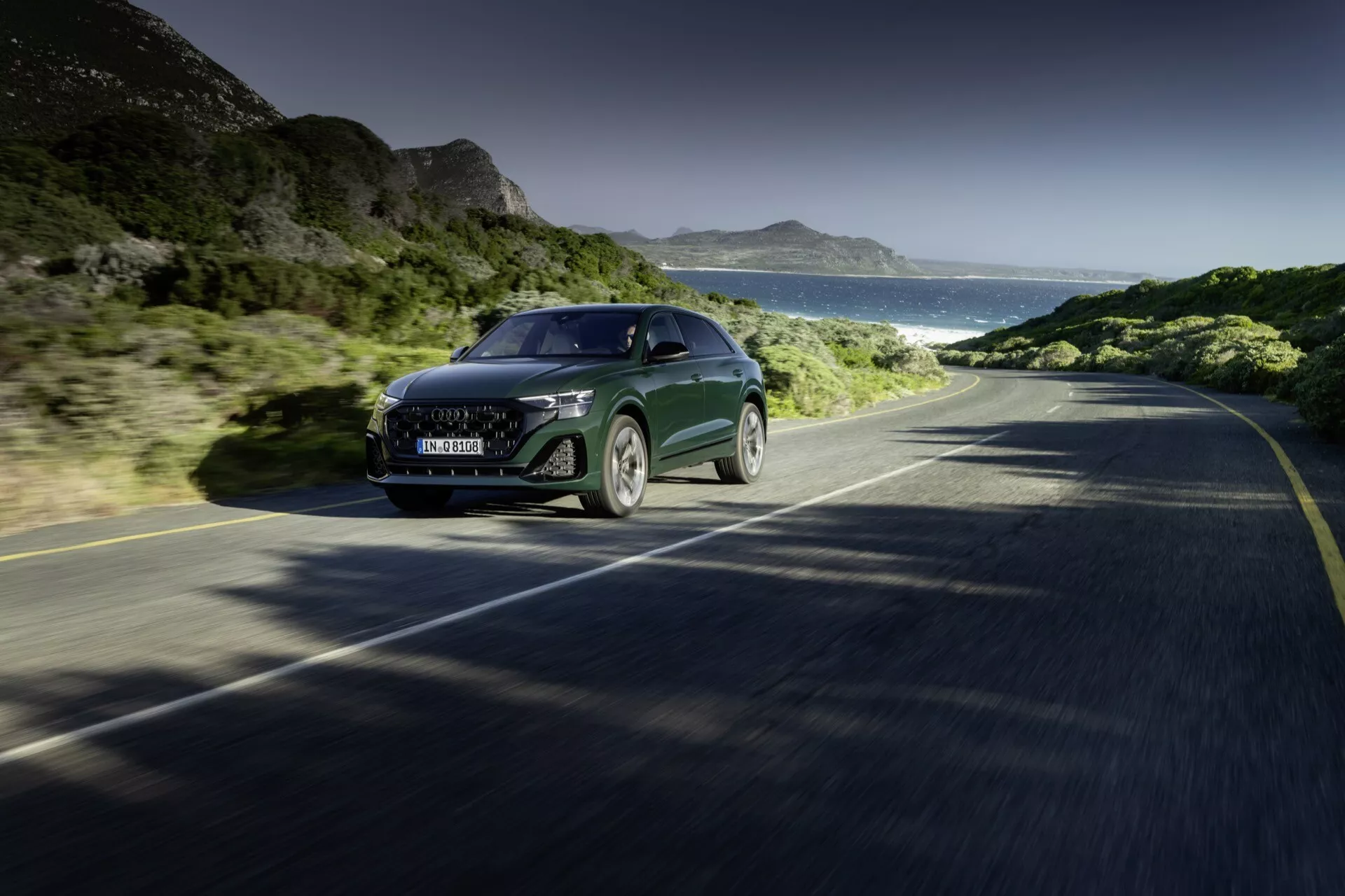 Audi Q8 2023 (4M-Facelift) vozi po obalni cesti, prikazuje pogled od spredaj-strani na njegov velik žar, LED žaromete, eleganten pokrov motorja in delno stransko ploščo, kar v gibanju ujame tehnologijo in luksuz proti ozadju jasnega neba, gora in oceana.