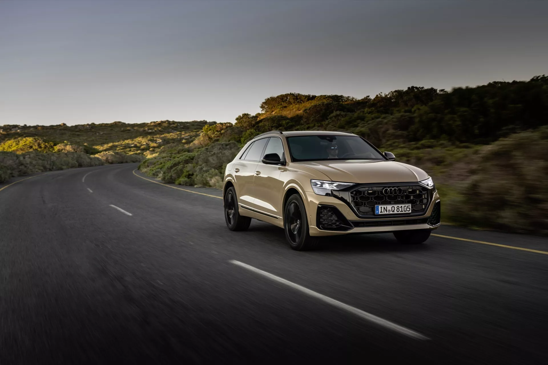 Audi Q8 2023 (4M-Facelift) v dinamični vožnji po ovinkasti cesti med zlatimi urami, prikazuje športno obliko kupeja avtomobila, agresivno sprednjo stran z LED žarometi in šesterokotno mrežo ter aerodinamično silhueto proti ozadju naravne pokrajine.