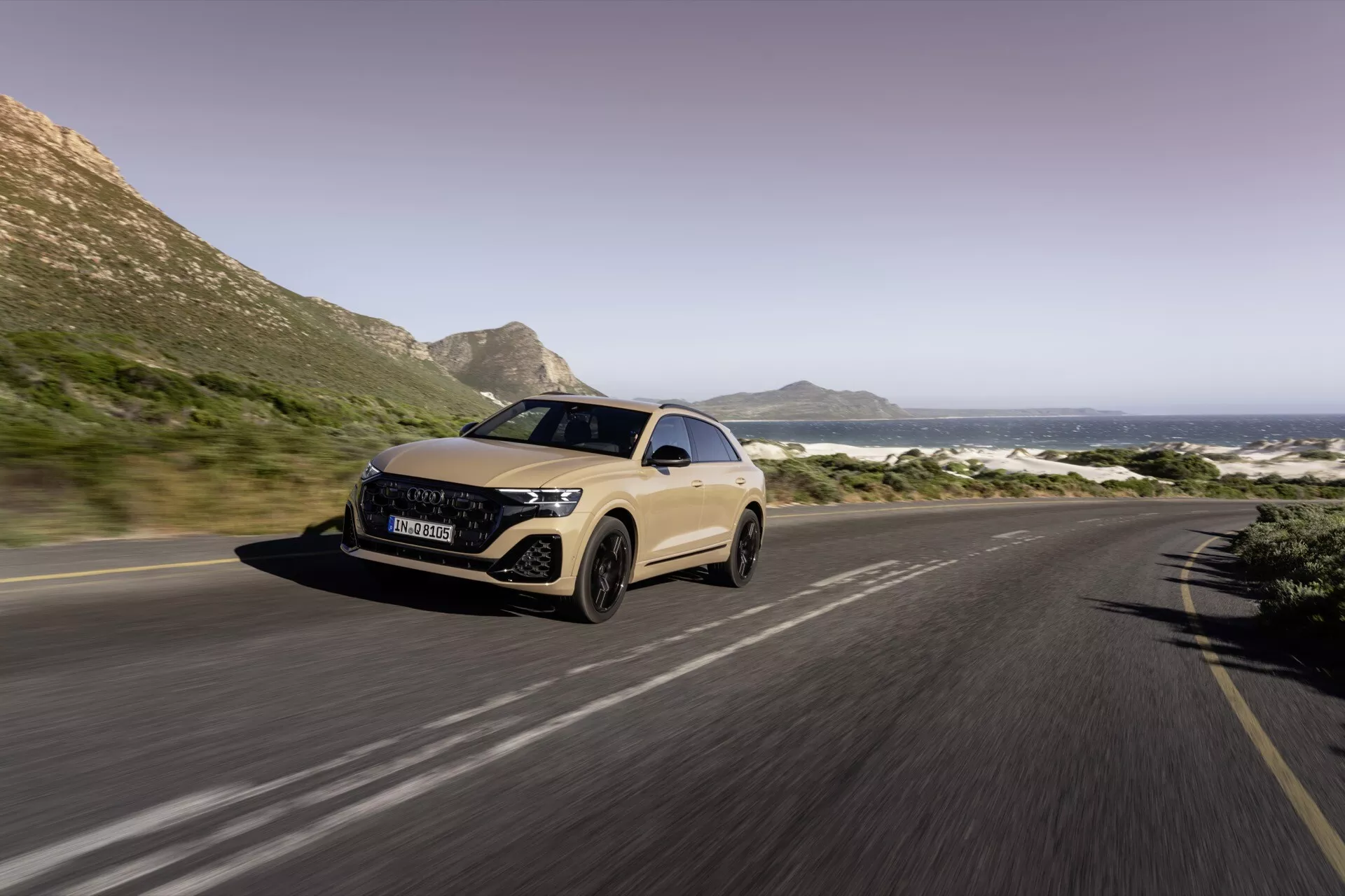 Tričetrtinski sprednji pogled na Audi Q8 2023 (4M-Facelift) na obalni cesti, ki prikazuje LED žaromete, aerodinamično zasnovo in zameglitev gibanja, ki kaže na premikanje