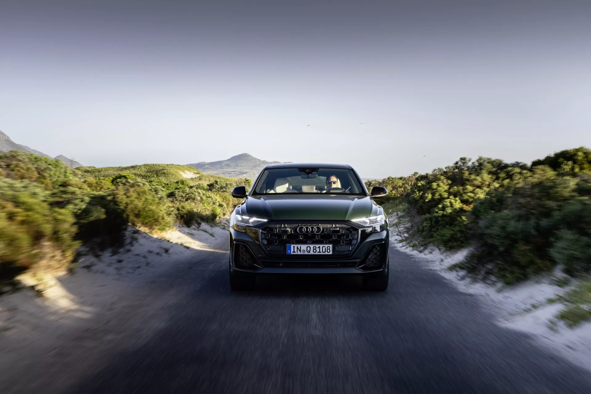 Audi Q8 4M-Facelift 2023 v gibanju, sprednji pogled prikazuje LED žarometi, napredno oblikovanje rešetke in aerodinamične lastnosti proti dinamičnemu ozadju ceste in gor