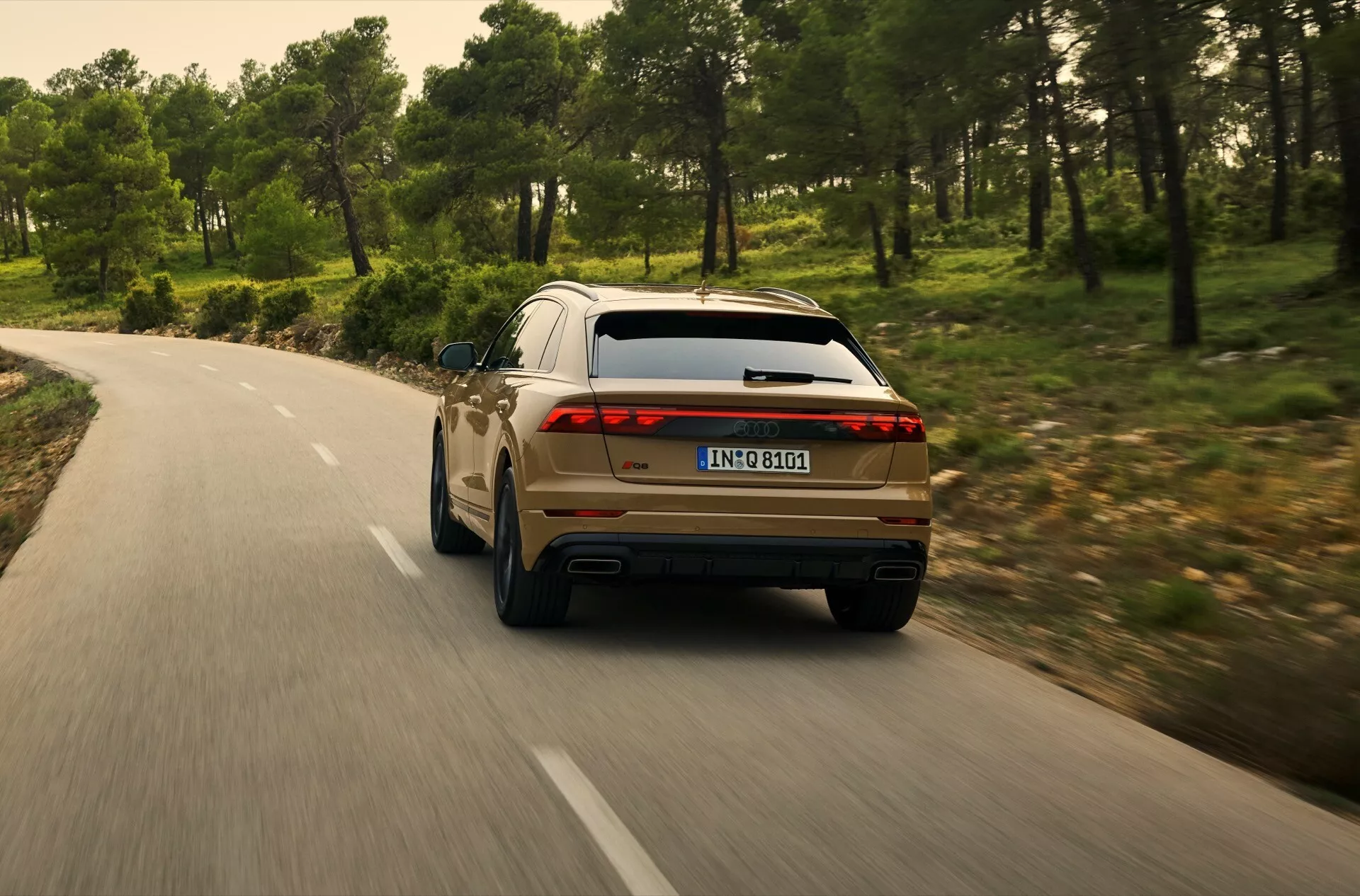 Zadnji pogled na Audi Q8 2023 (4M-Facelift), ki vozi po ovinkasti cesti skozi gozd, prikazuje desno zadnje četrtino z LED zadnjimi lučmi, dvojnim izpuhom in sodobnimi oblikovalskimi značilnostmi