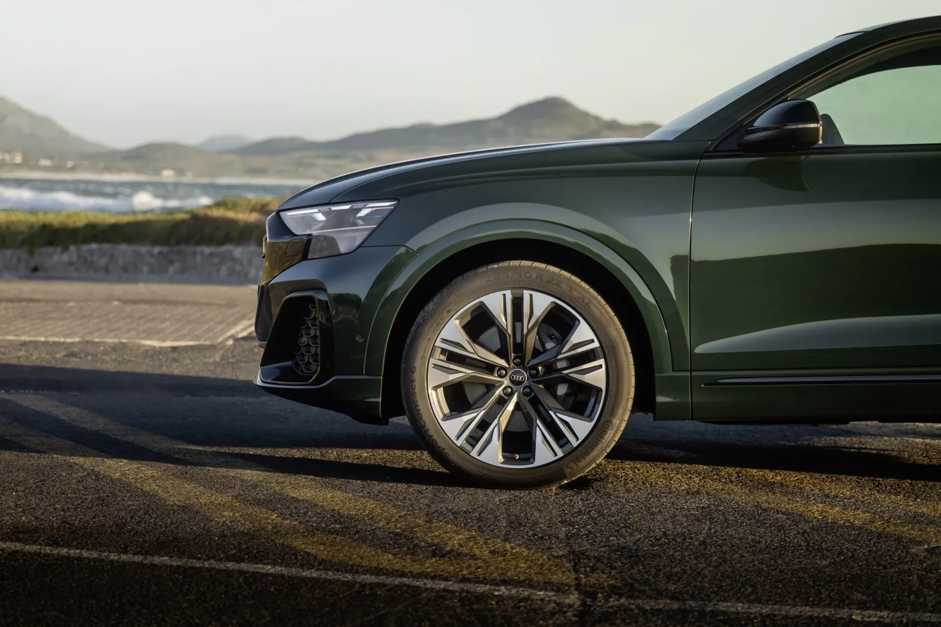 Pogled od blizu na športni sprednji levi del Audi Q8 2023 (4M-Facelift), vključno s poliranim večkrakim zlitinskim kolesom, delnim sprednjim odbijačem z velikim zračnim vhodom, robom sodobne LED žaromete in sijajno globoko zeleno metalik barvo na karoseriji, proti zunanji kulisi.