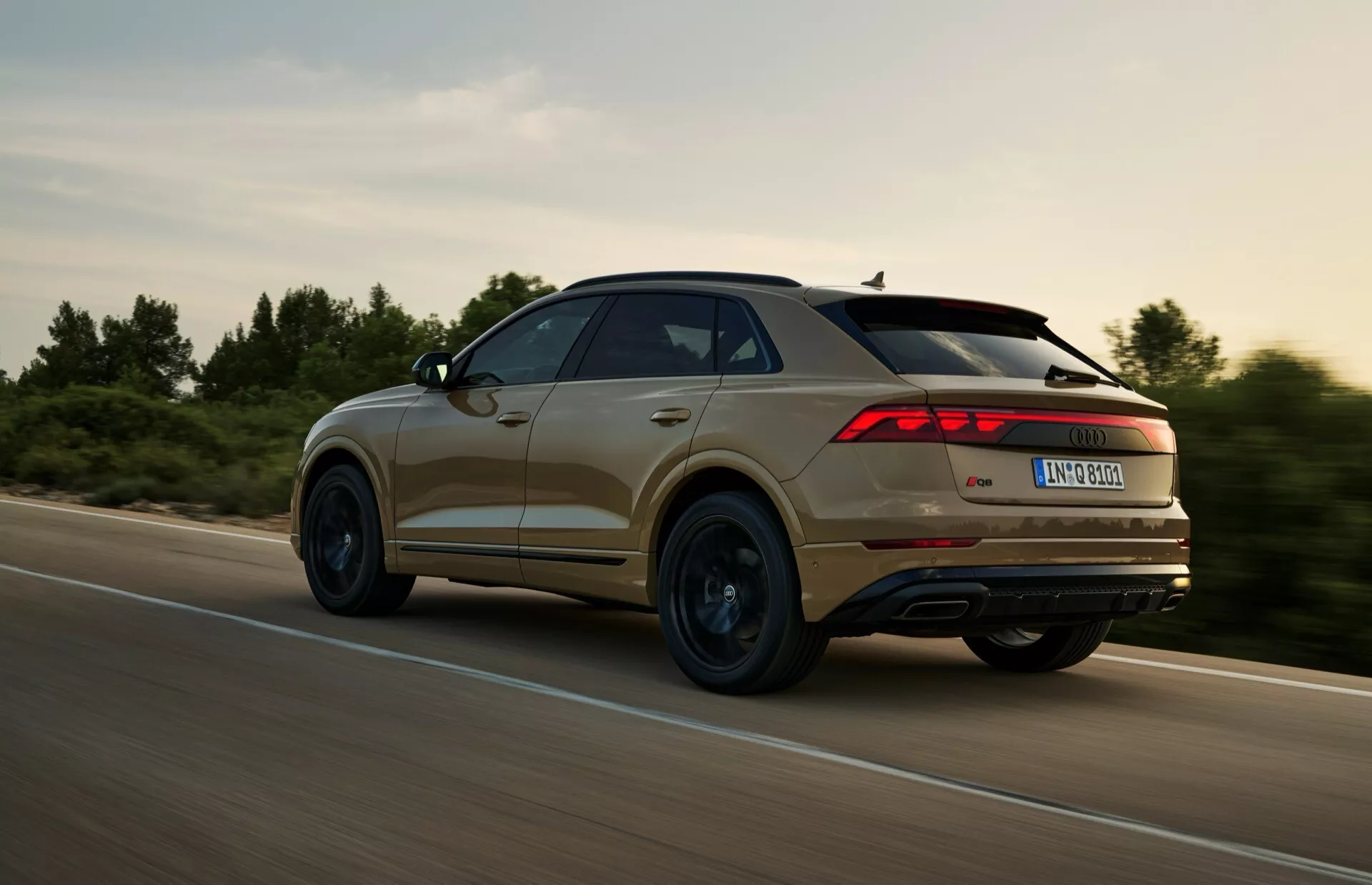 Zadnji tričetrtinski pogled na SUV Audi Q8 (4M-Facelift) letnik 2023 v gibanju, z izrazitim dizajnom zadnjih luči, konturnimi linijami in profilom kolesa, posnet med dinamično vožnjo v podeželskem okolju s toplo razsvetljavo.