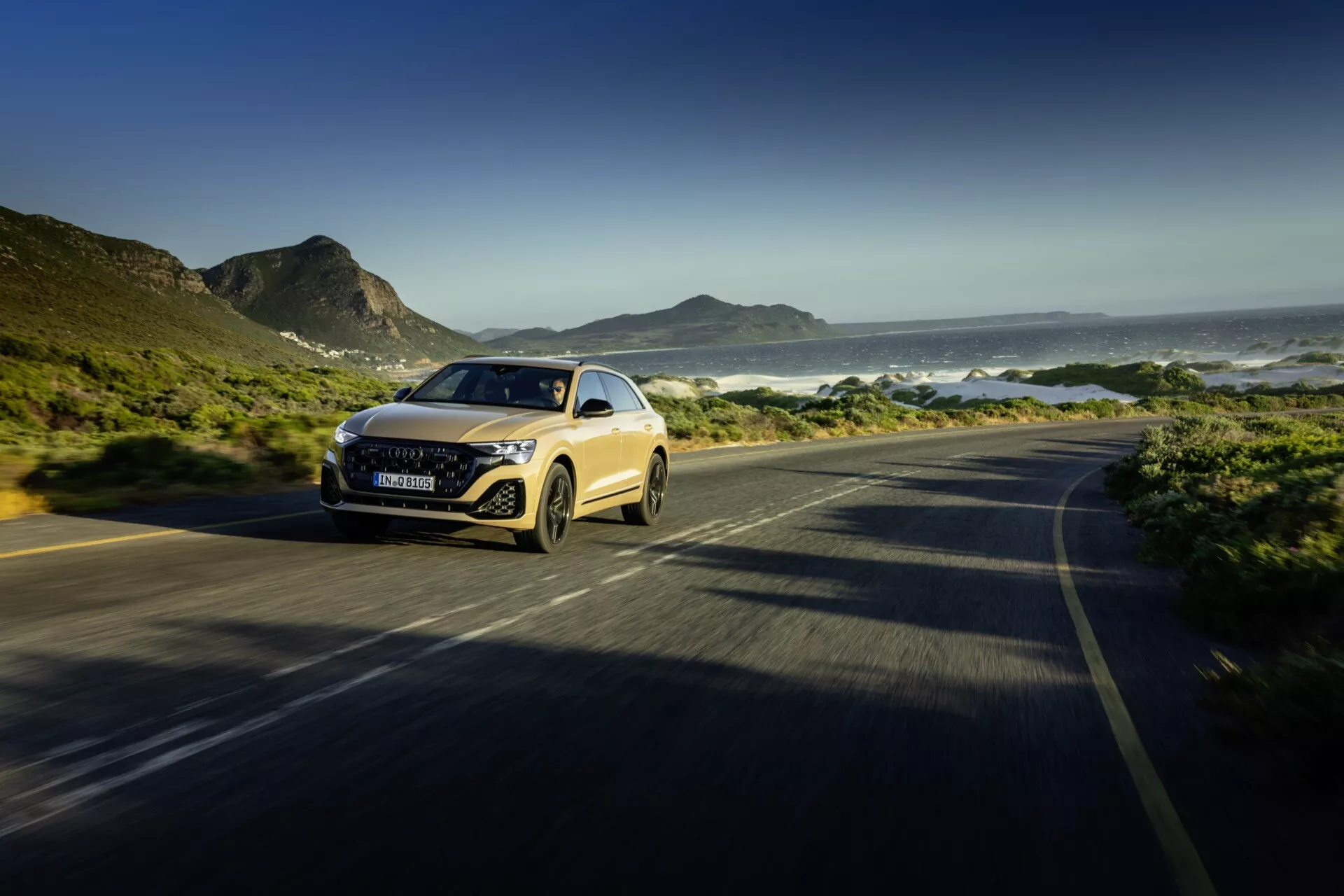 Audi Q8 2023 (4M-Facelift) vozi po obalni cesti, kar prikazuje njegov dinamični dizajn in LED žaromete v pogledu spredaj tri četrtine z zamegljenim gibanjem