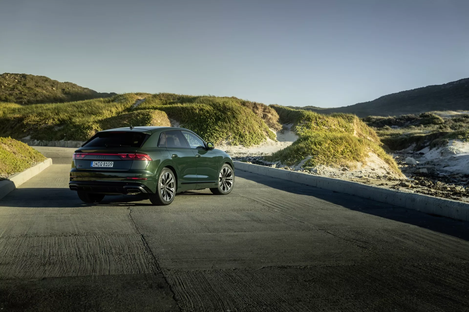 Audi Q8 4M-Facelift 2023 prikazan proti obalni pokrajini sipin v zlati uri, prikazuje pogled s treh četrtin zadaj z modernimi vodoravnimi zadnjimi lučmi in elegantno zasnovo