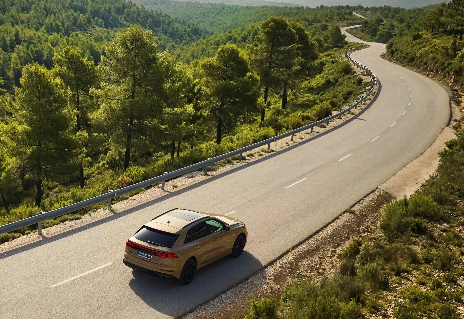 Zlato Audi Q8 2023 (4M-Facelift) na ovinkasti dvojni cesti v ruralnem ali gozdnem okolju, pogled iz povišanega kota, ki prikazuje njegov zadnji in stranski dizajn proti naravni krajini