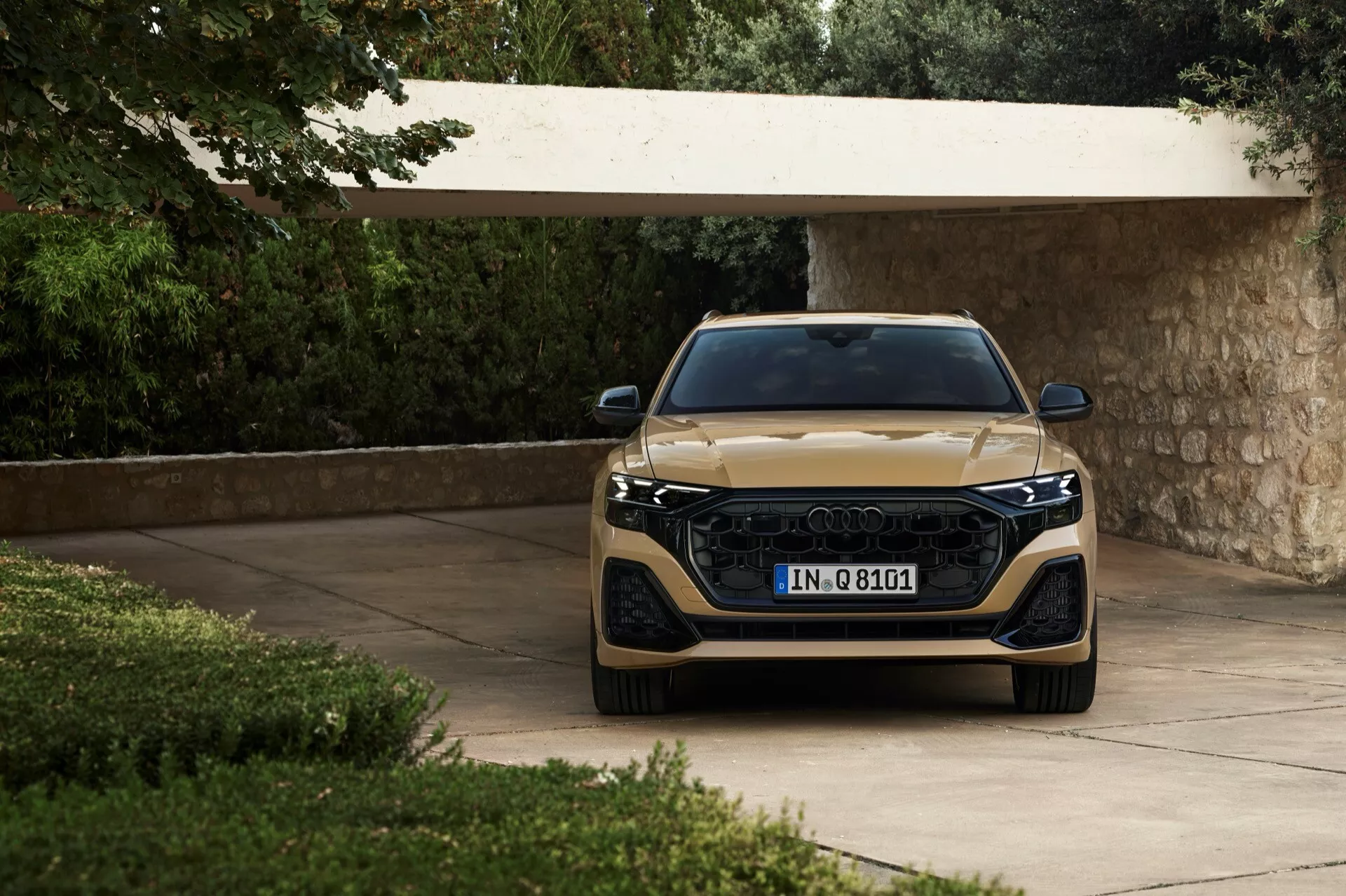 Audi Q8 2023 (4M-facelift) prikazan iz frontalnega kota, poudarja njegov širok žar, elegantne LED žaromete in kovinski zaključek, postavljen na dovozni poti, obdani z bujnim zelenjem