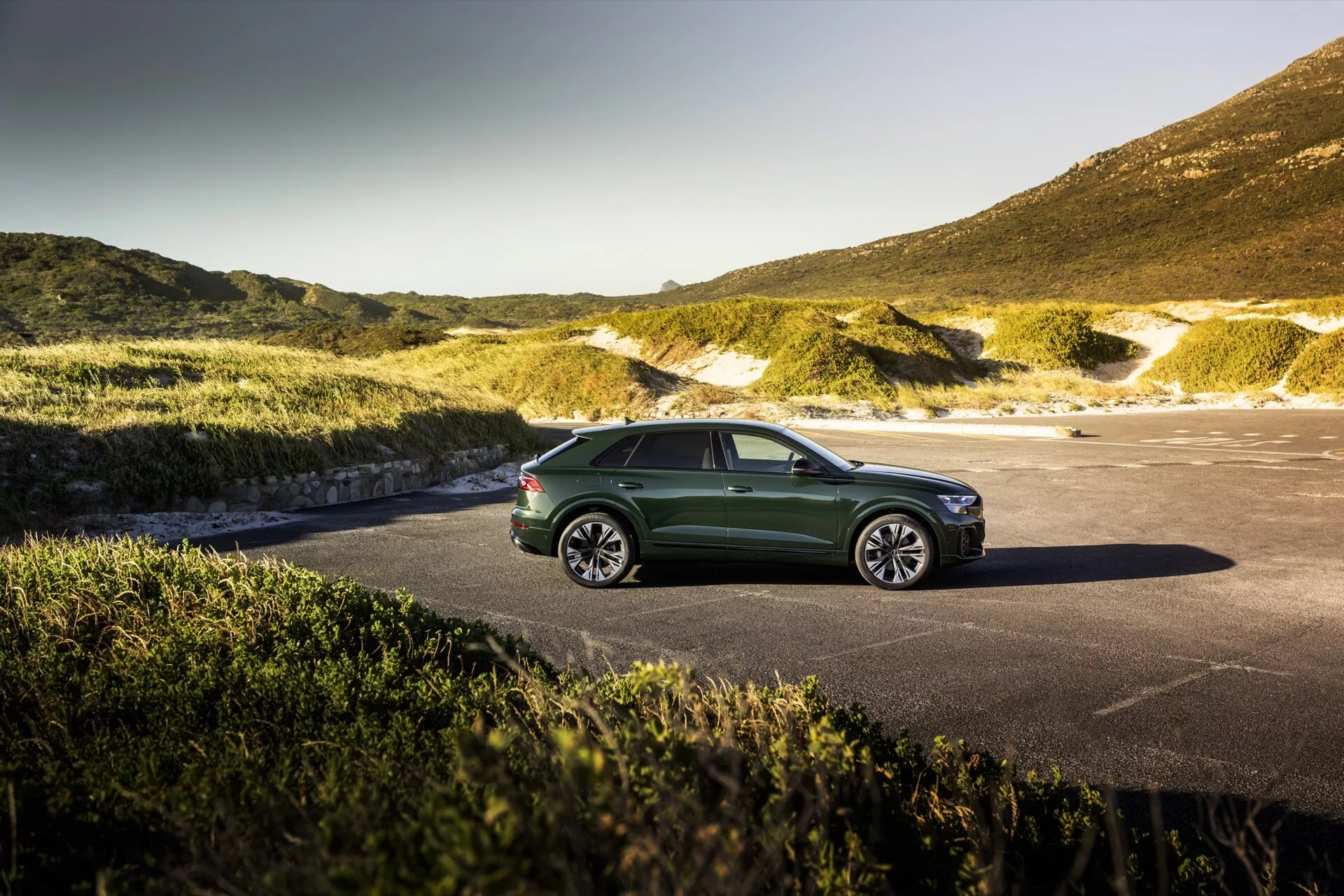 Audi Q8 2023 (4M-Facelift) v zeleni parkiran s svojo elegantno aerodinamično zasnovo in LED žarometi proti slikovitem naravnem ozadju, morda obalnem ali gorskem, pod jasnim nebom