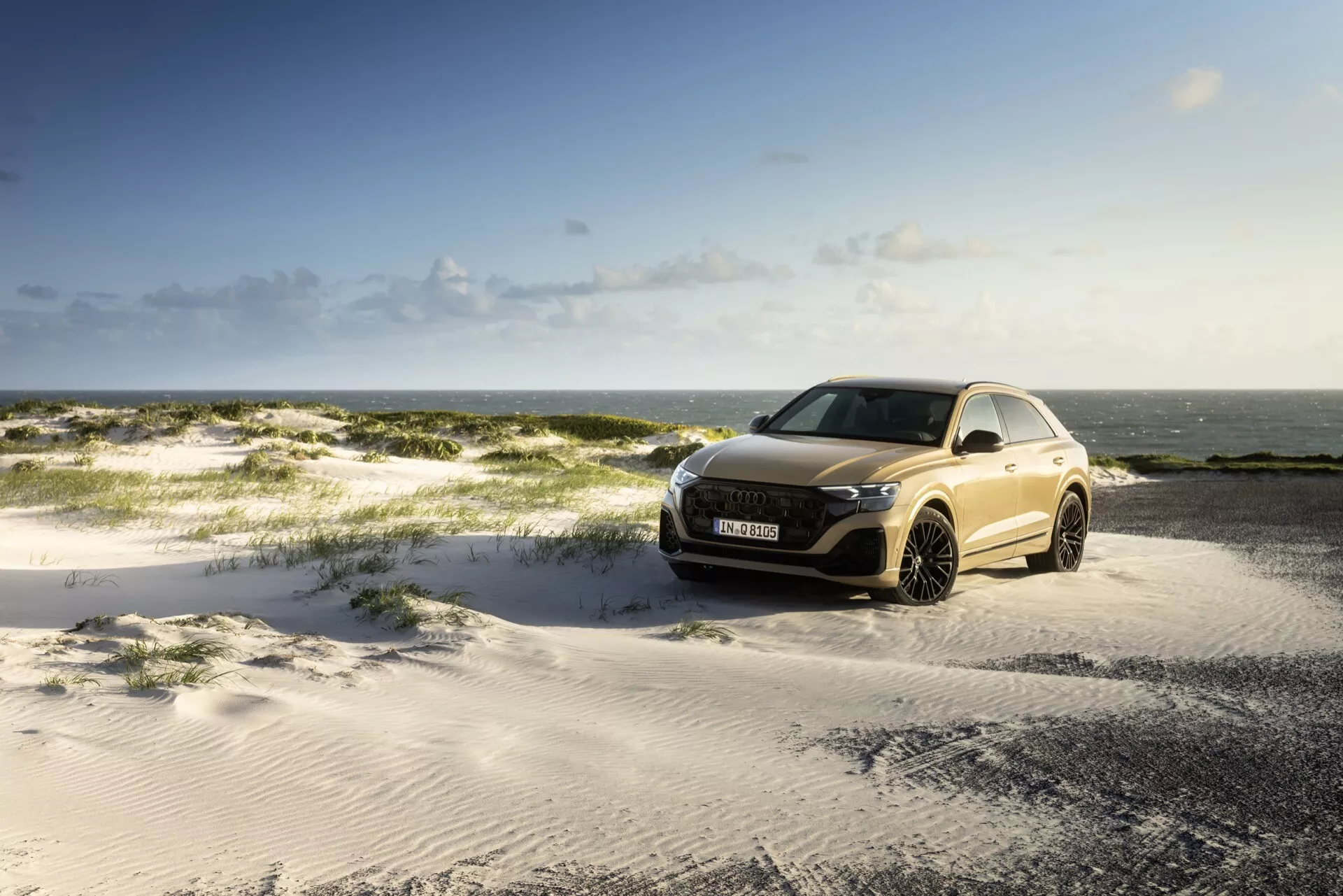 Audi Q8 2023 (4M-Facelift) parkiran na peščenem terenu pod delno oblačnim nebom z morjem v ozadju, ki prikazuje sprednjo rešetko, LED žaromete in aerodinamično zasnovo